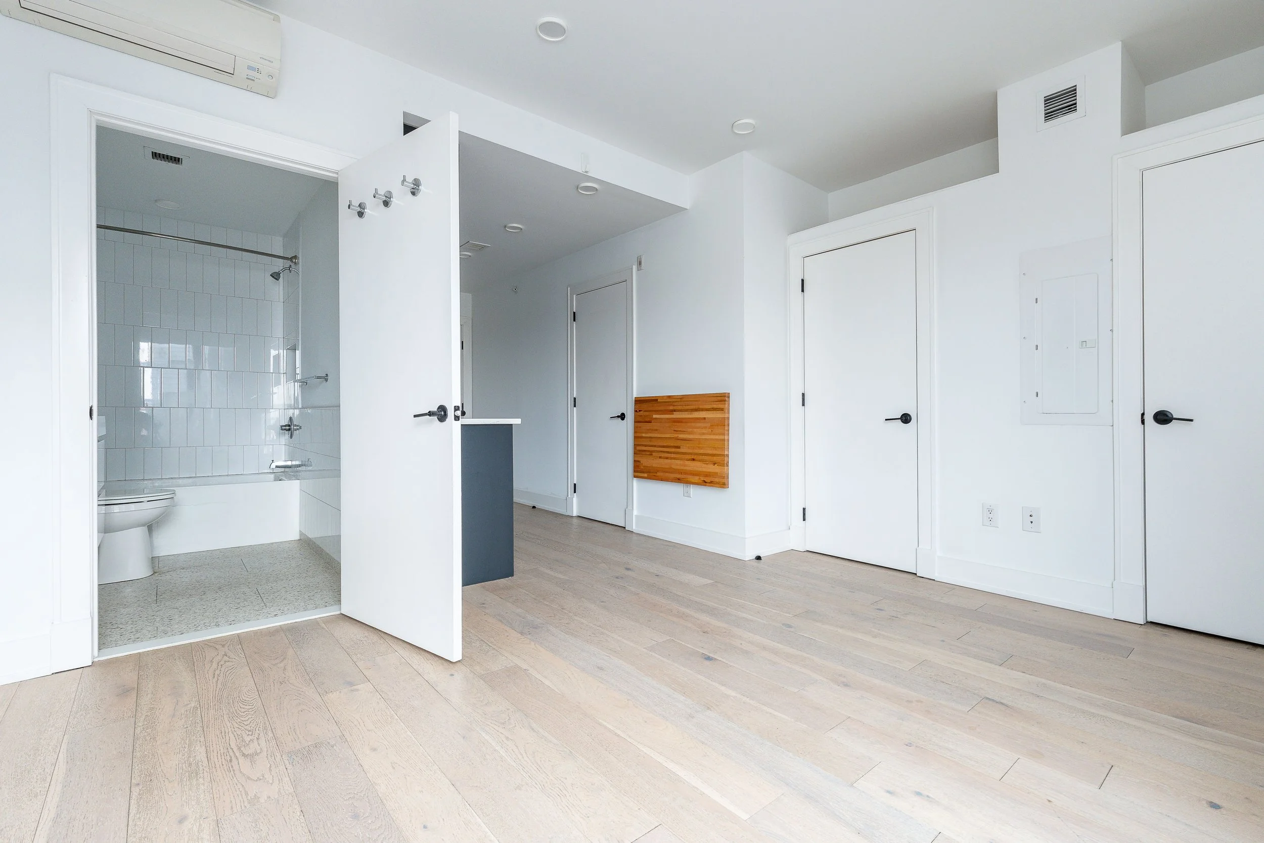 Empty room with light wood flooring, white walls, and a doorway leading to a bathroom with white tiled shower area and toilet. An air conditioning unit is mounted on the wall, and there are multiple closed white doors in the room.