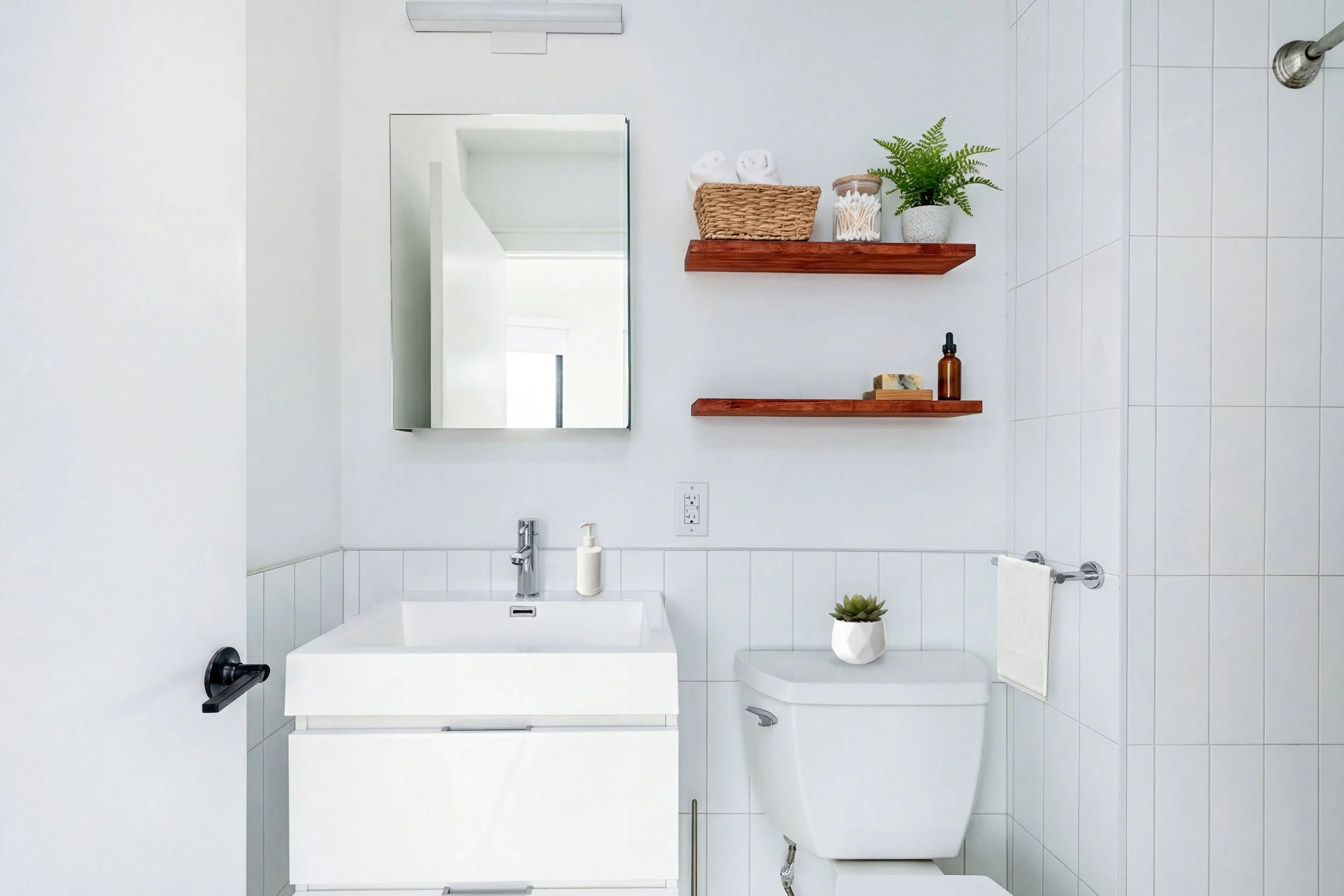 Minimalist bathroom with a floating vanity, a mirror, two floating wooden shelves with decor, and a white toilet with a potted plant on top.
