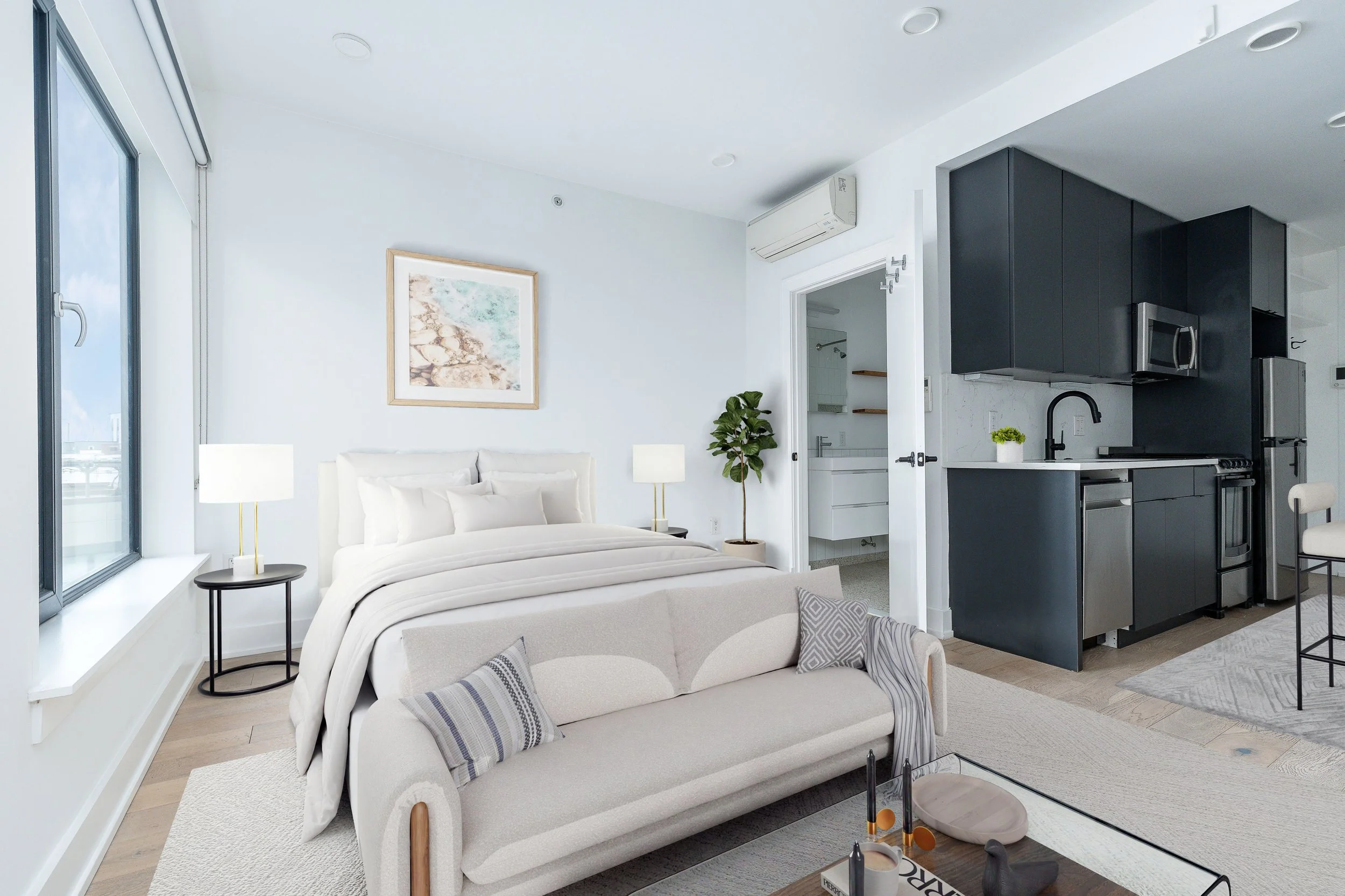 Modern studio apartment featuring a sleeping area with a bed, a living area with a sofa, a small tabletop with decor, and a kitchen with dark cabinets and stainless steel appliances.