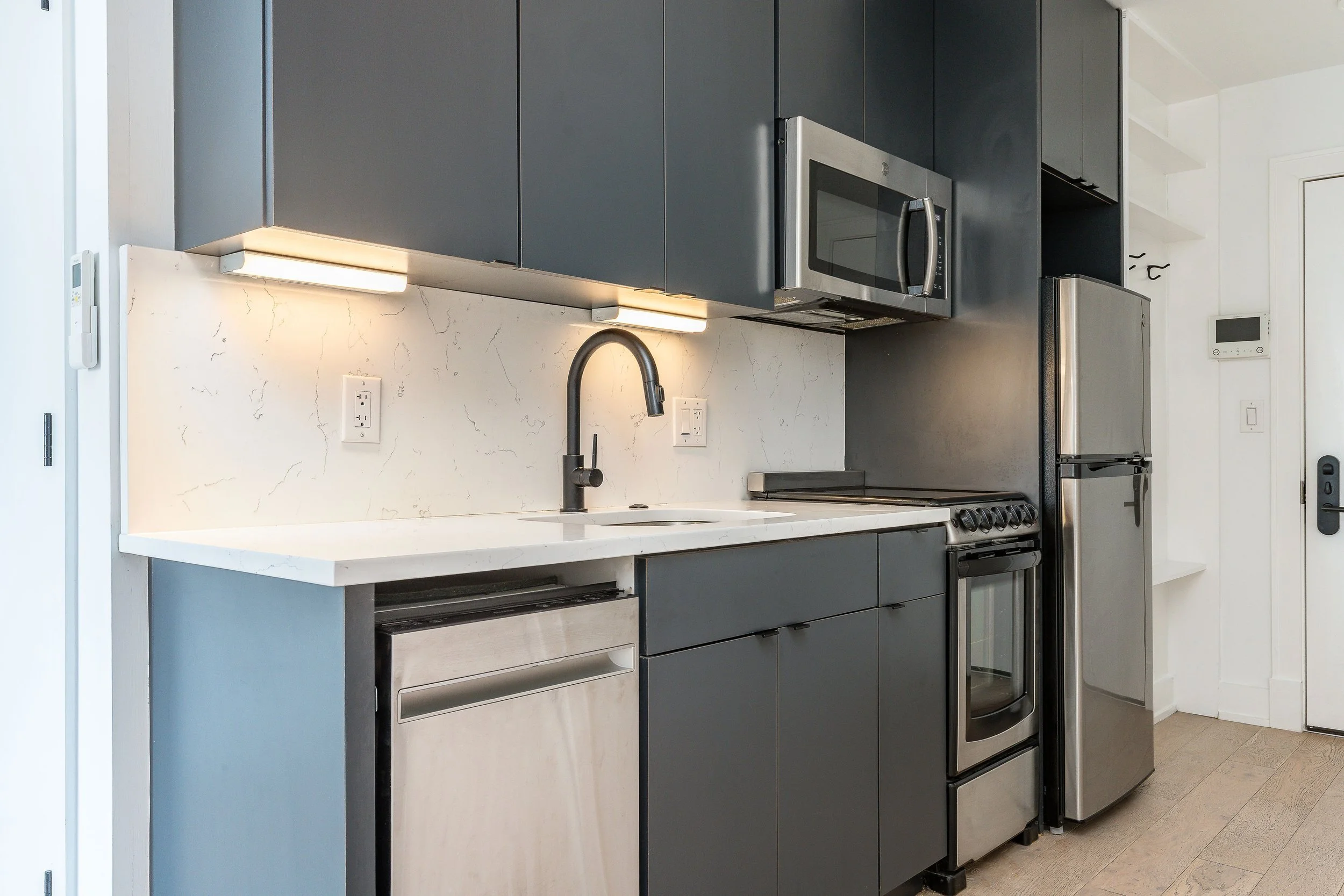 Modern kitchen with dark gray cabinets, white marble countertop, stainless steel appliances including refrigerator, oven, microwave, and dishwasher, and a black faucet at the sink.
