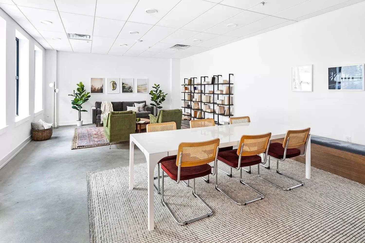 A modern office lounge featuring a seating area with a black sofa, green armchairs, and a bookshelf against a white wall. A dining area with a white table and six chairs is in the foreground.
