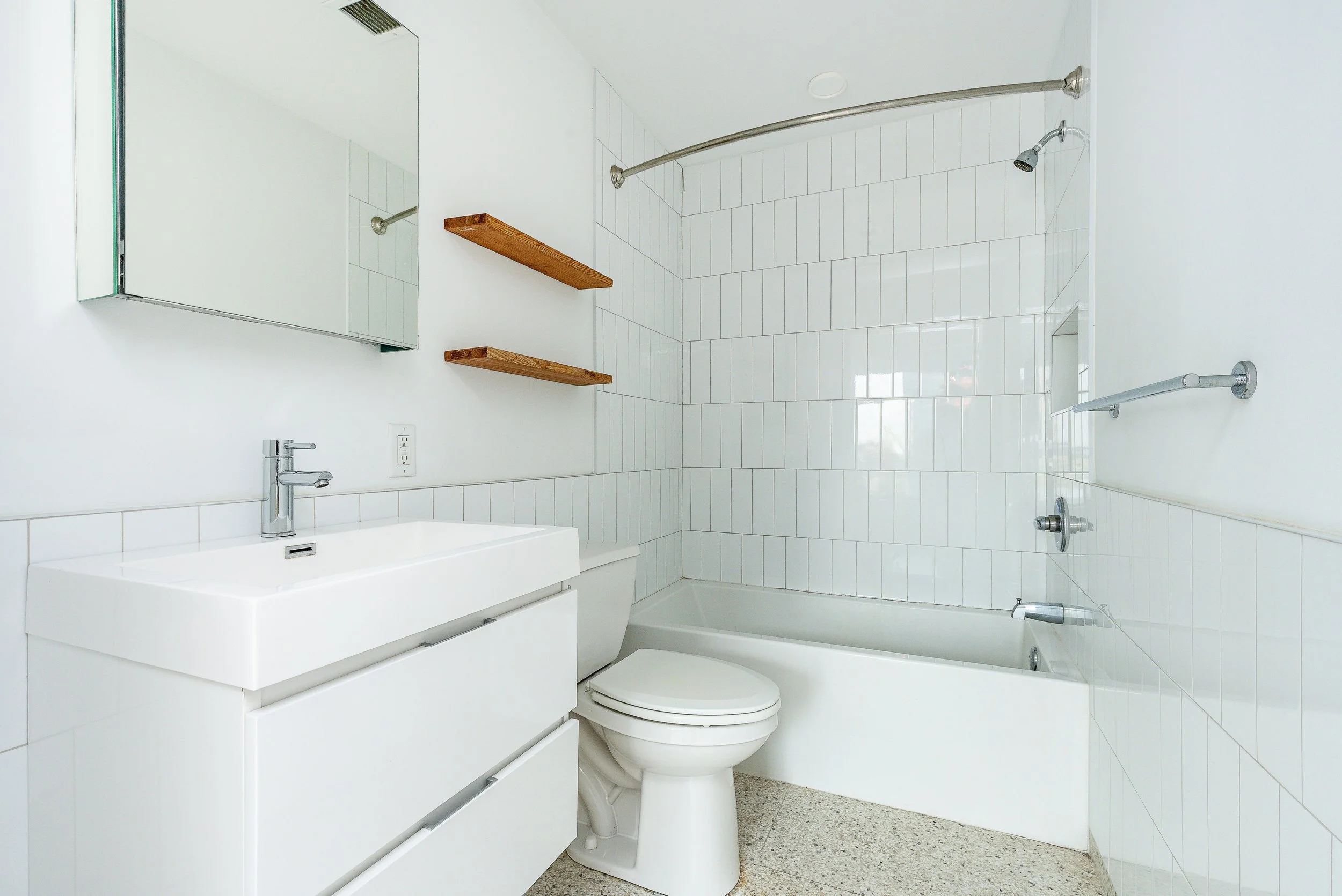 A minimalist white bathroom with a white vanity, toilet, and bathtub. The wall has white tiles and two wooden shelves. The bathroom features a mirror cabinet above the sink and silver fixtures.