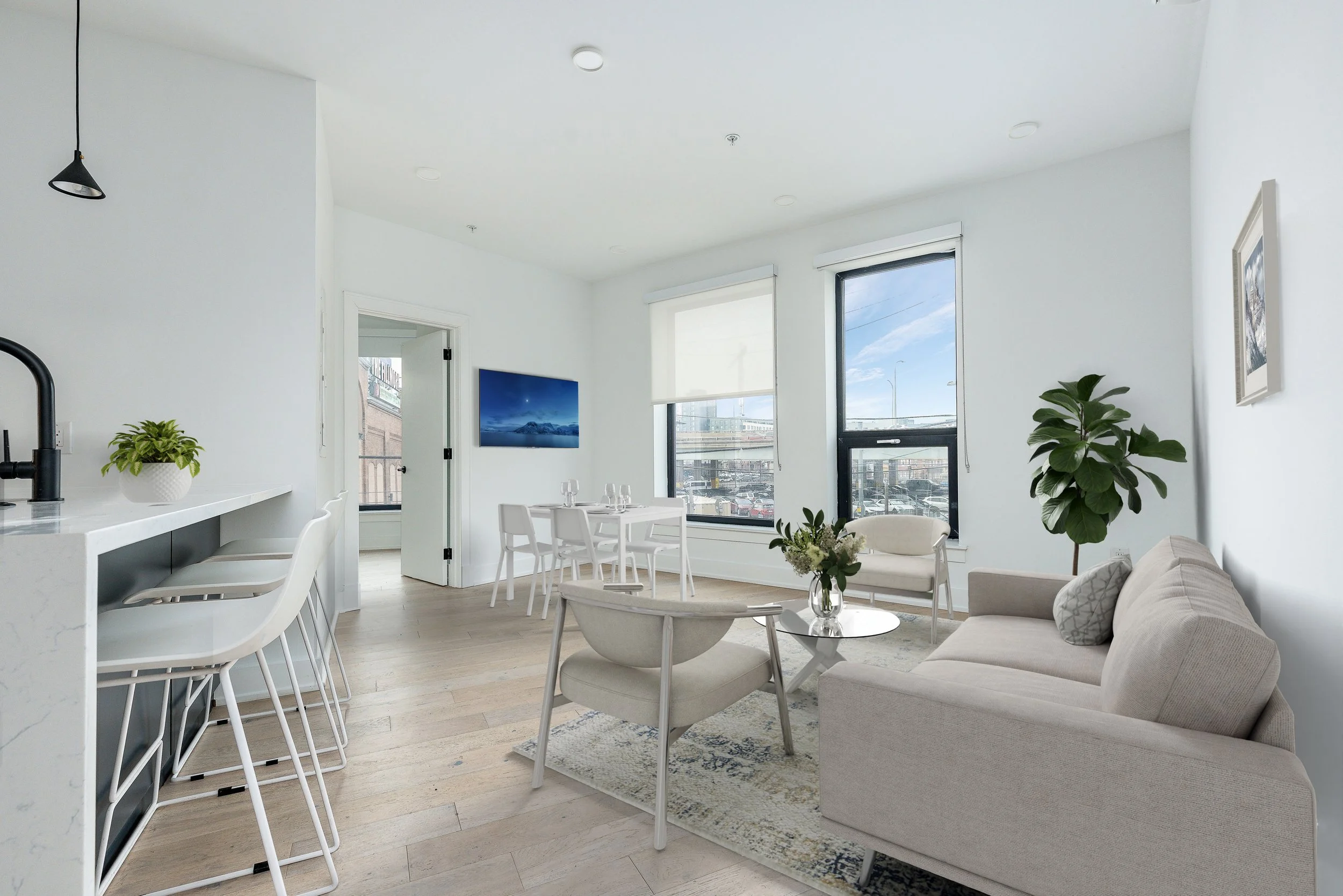 Bright modern living room with large windows, white walls, beige furniture, a small white armchair, and a beige sofa, with a cream-colored rug, potted plants, and wall art.