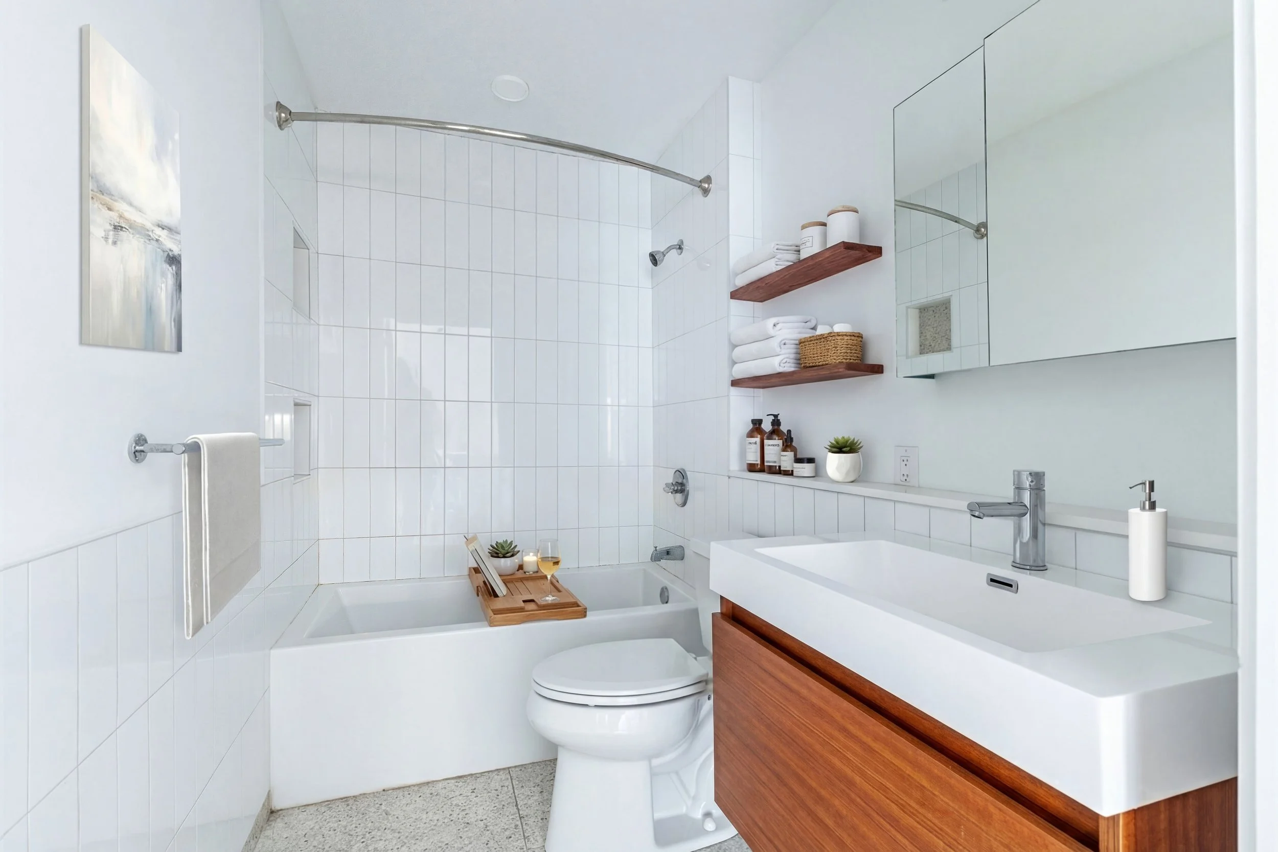 Modern bathroom with white tiled walls, a bathtub with a wooden tray holding a wine glass and succulent, a white toilet, a wooden vanity with a white sink, wall-mounted shelves with towels, toiletries, a potted plant, and a mirrored cabinet.