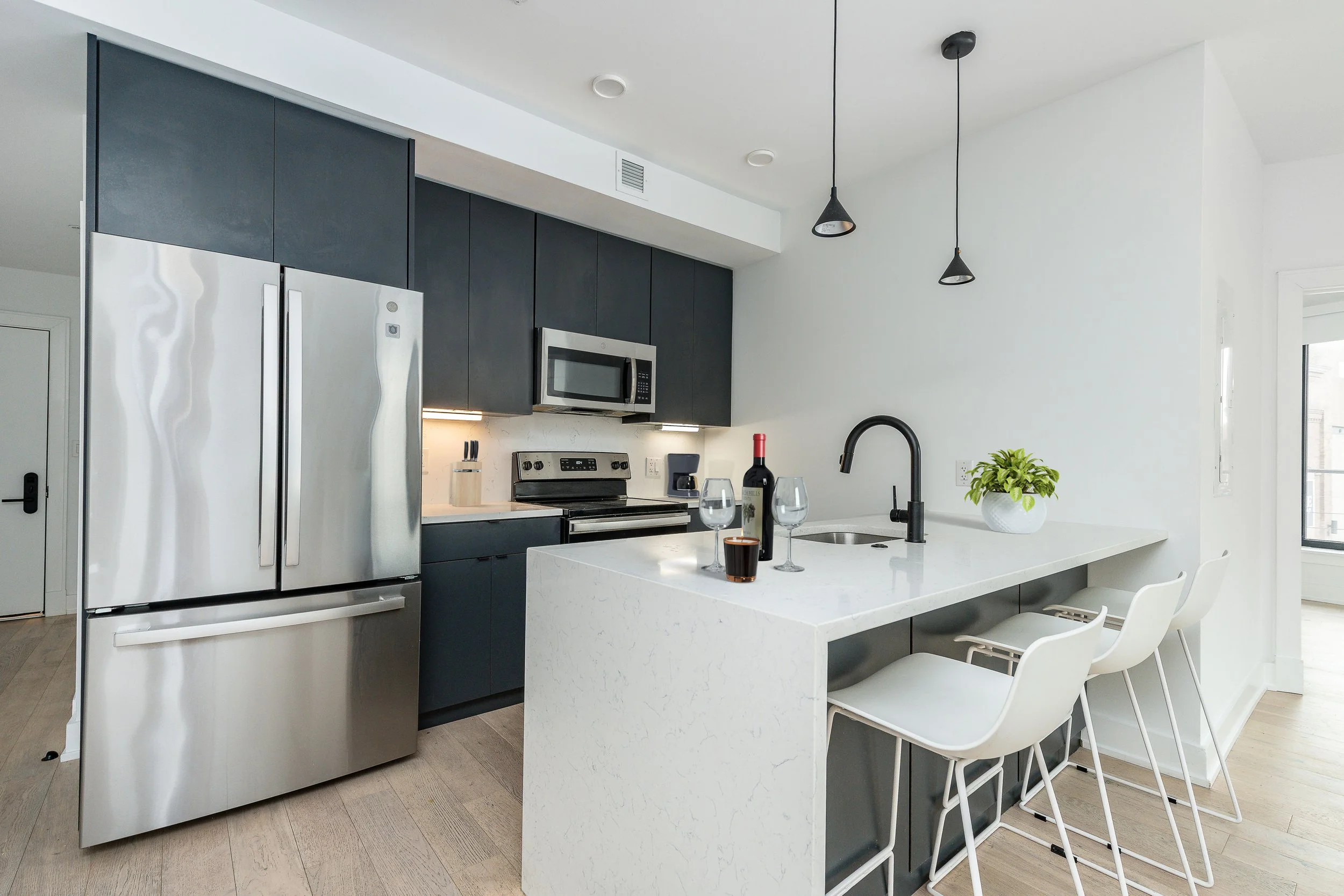 Modern kitchen with stainless steel refrigerator, black cabinetry, white countertops, and bar stools. Items include a wine bottle, glasses, and a coffee.