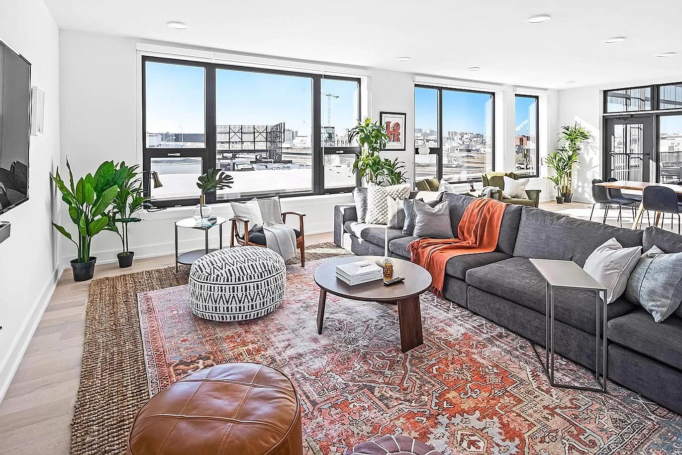 Modern living room with large windows, gray sectional sofa, round coffee table, patterned ottoman, leather pouf, plants, and colorful rugs.