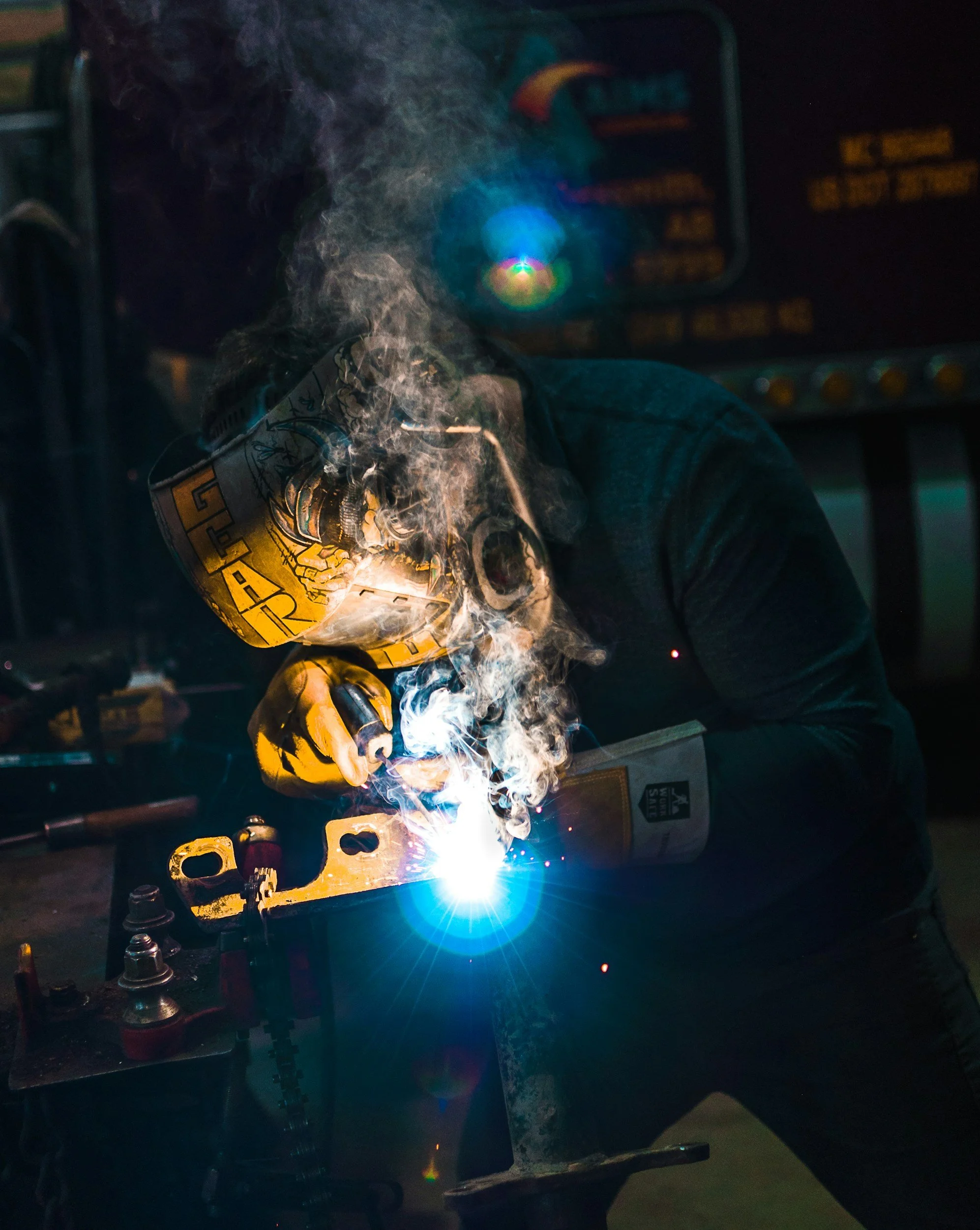 Una persona sujetando con guantes amarillos y negras un motor de motosierra, mientras realiza una soldadura que emite chispas y humo en un ambiente oscuro. En el fondo, hay una pantalla con controles y un panel de instrumentos.