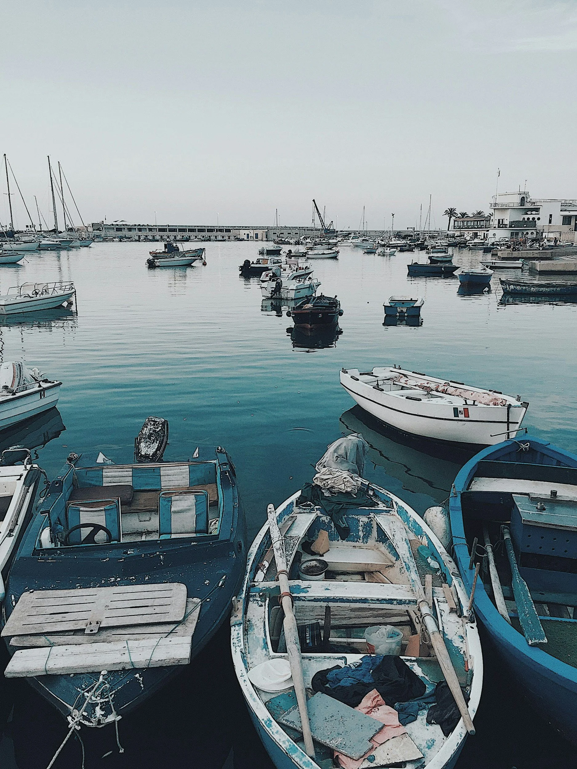 Barco de vela y otras embarcaciones en el puerto, con un muelle y edificios en el fondo.