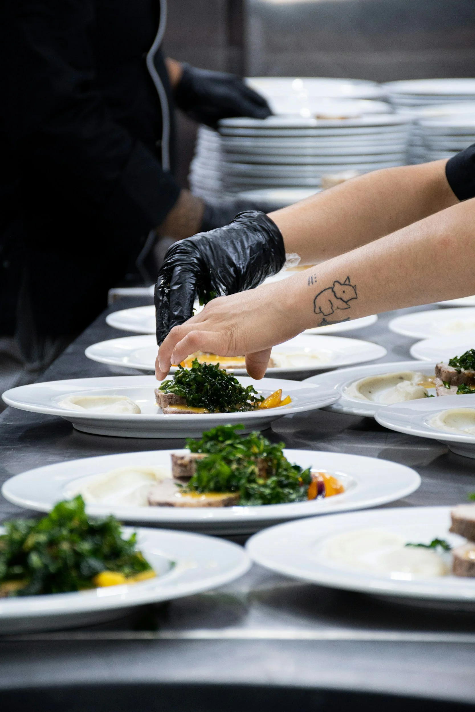 Personas sirviendo platos con comida en un entorno de cocina profesional o evento de servicio de alimentos, con platos blancos y comida decorativa.