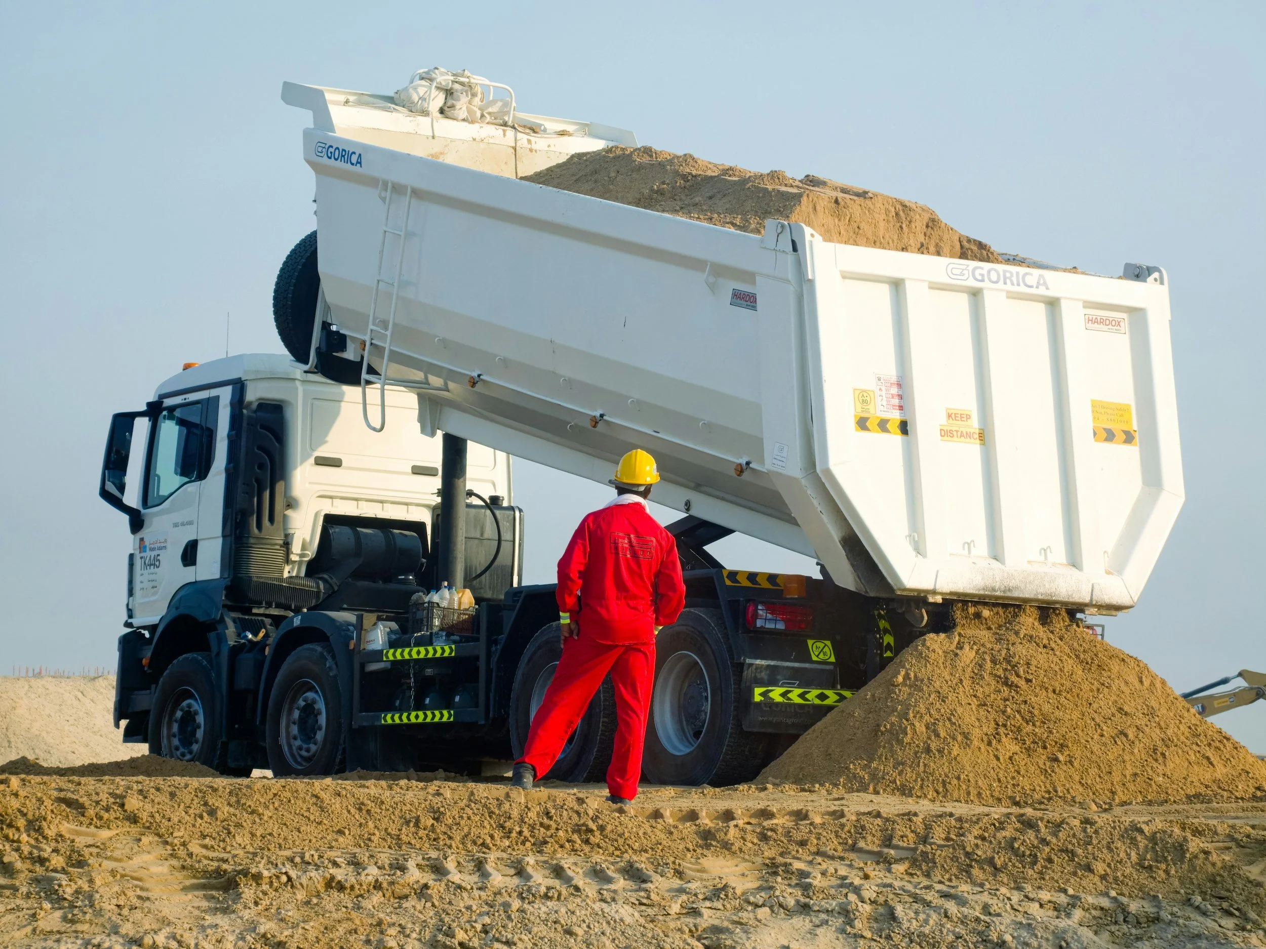 Vehículo de construcción con una grúa descargando arena o tierra en un sitio de obra, con un trabajador con uniforme rojo y casco amarillo observando.