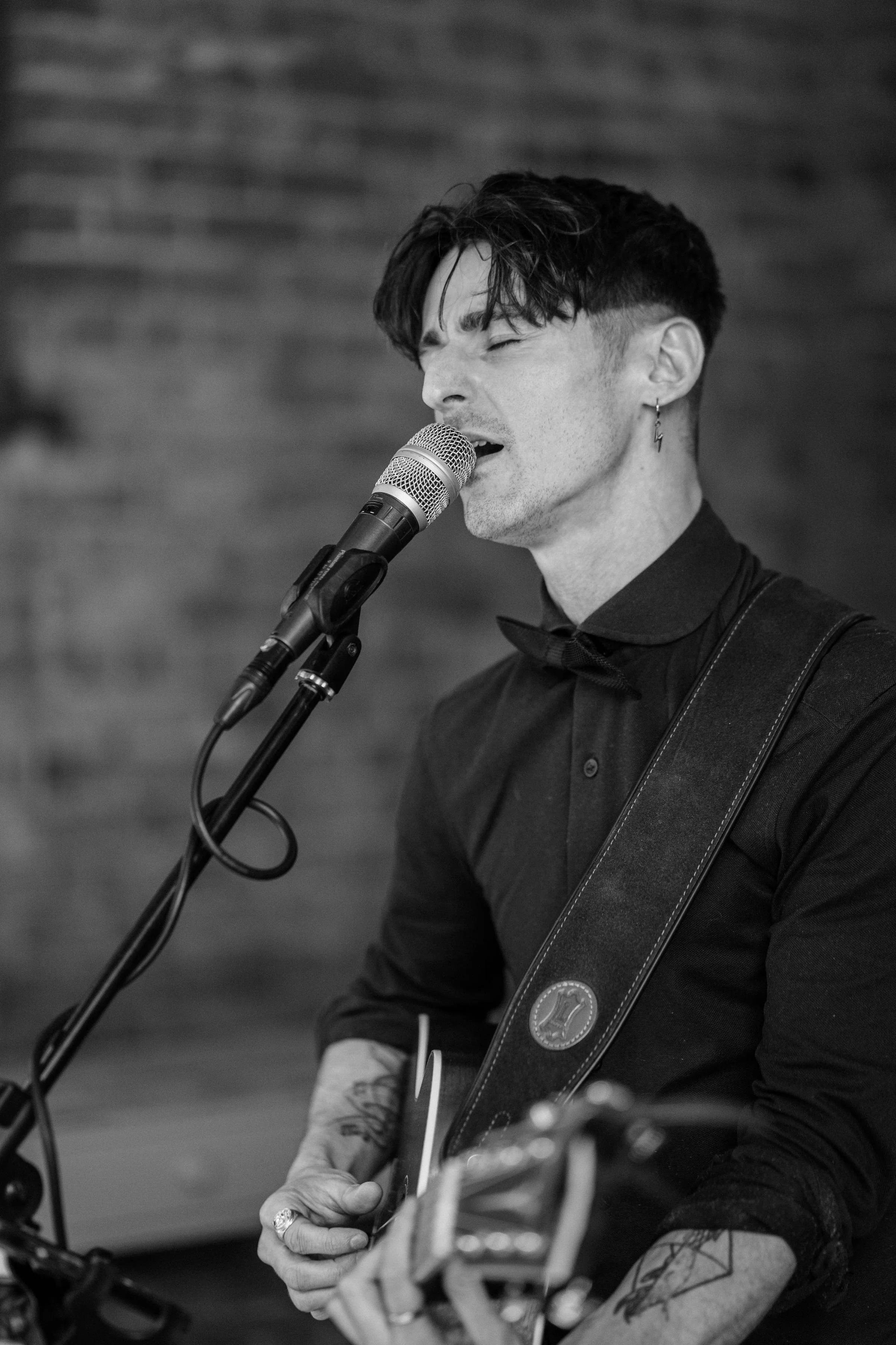 A male wedding musician singing into a microphone while playing an acoustic guitar, wearing a black shirt and a leather strap, with tattoos and an earring, performing in front of a brick wall.