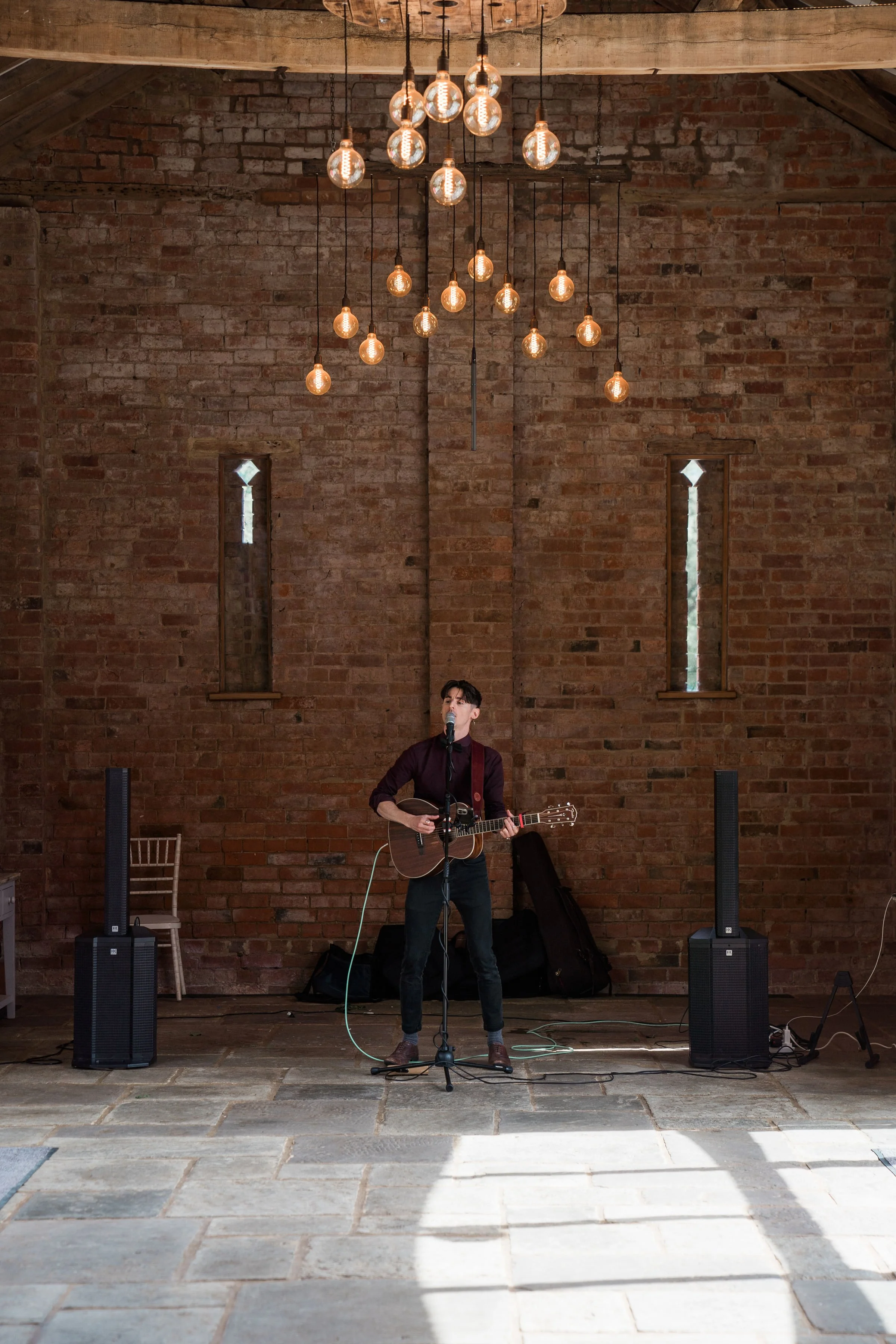 A man singing and playing guitar on a stage at a wedding with brick wall background, hanging light bulbs, and a microphone. There are two tall speakers on either side of him.