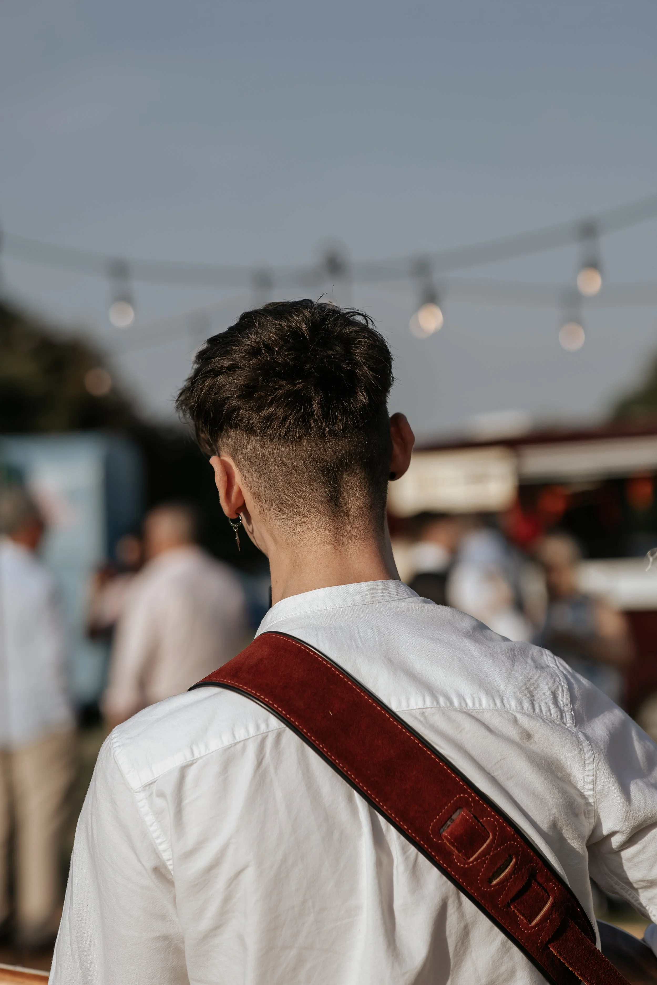 Back of a person with short, dark hair, wearing a white shirt and a brown, leather strap over their shoulder . Blurred people and wedding lights in the background outdoors.