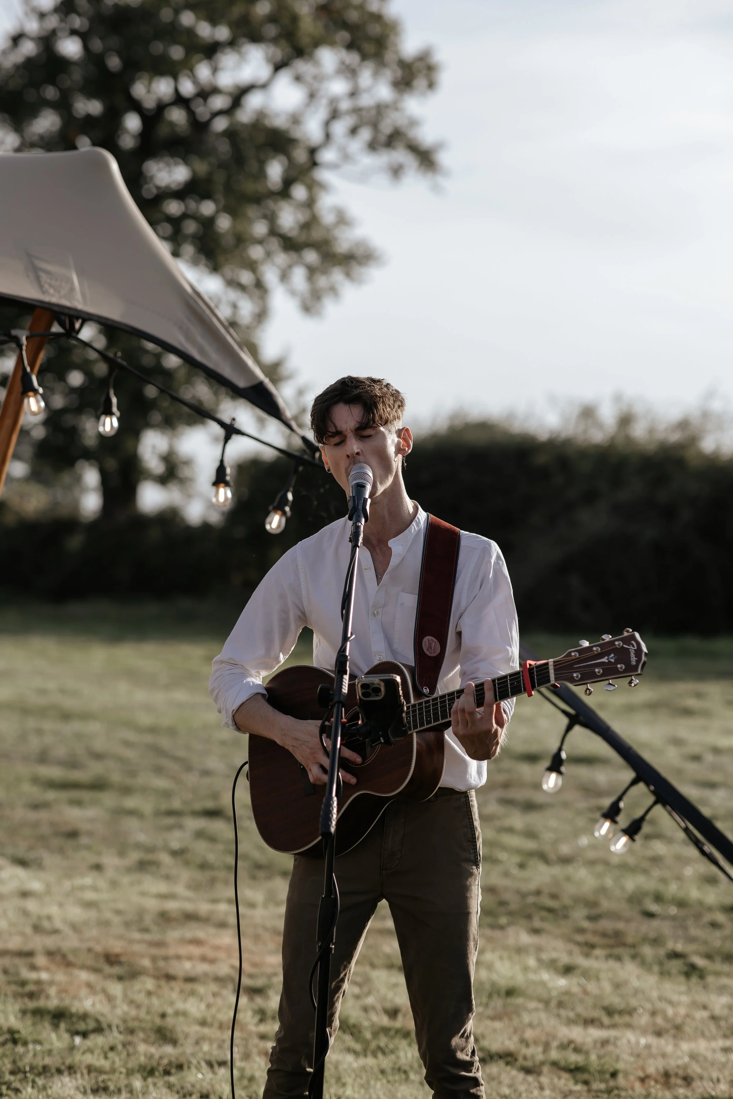 A young man sings and plays guitar outdoors during a wedding performance, with string lights hanging nearby and trees in the background.