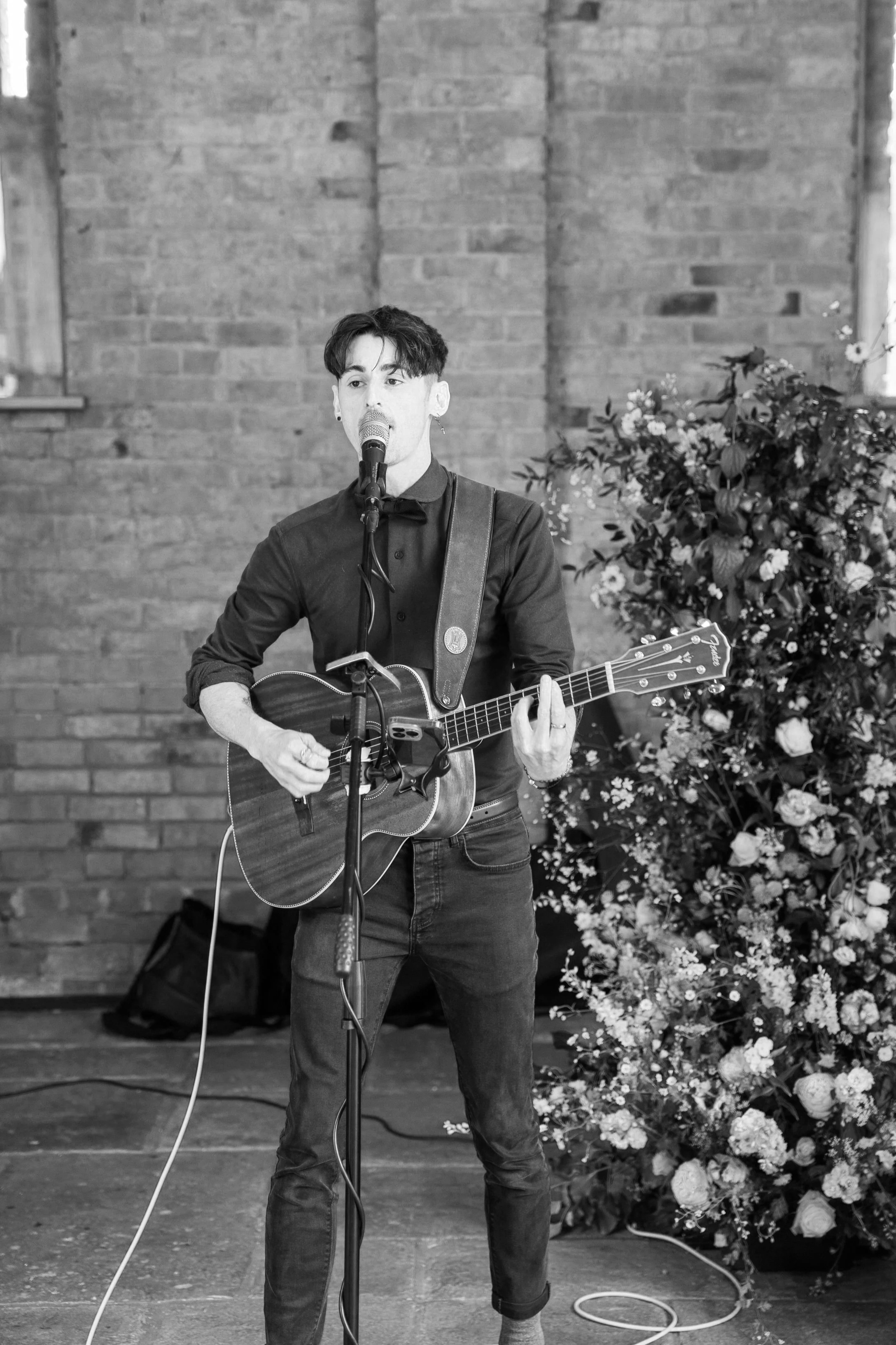 A man singing into a microphone and playing an acoustic guitar during a wedding performance, with a large floral arrangement in the background.