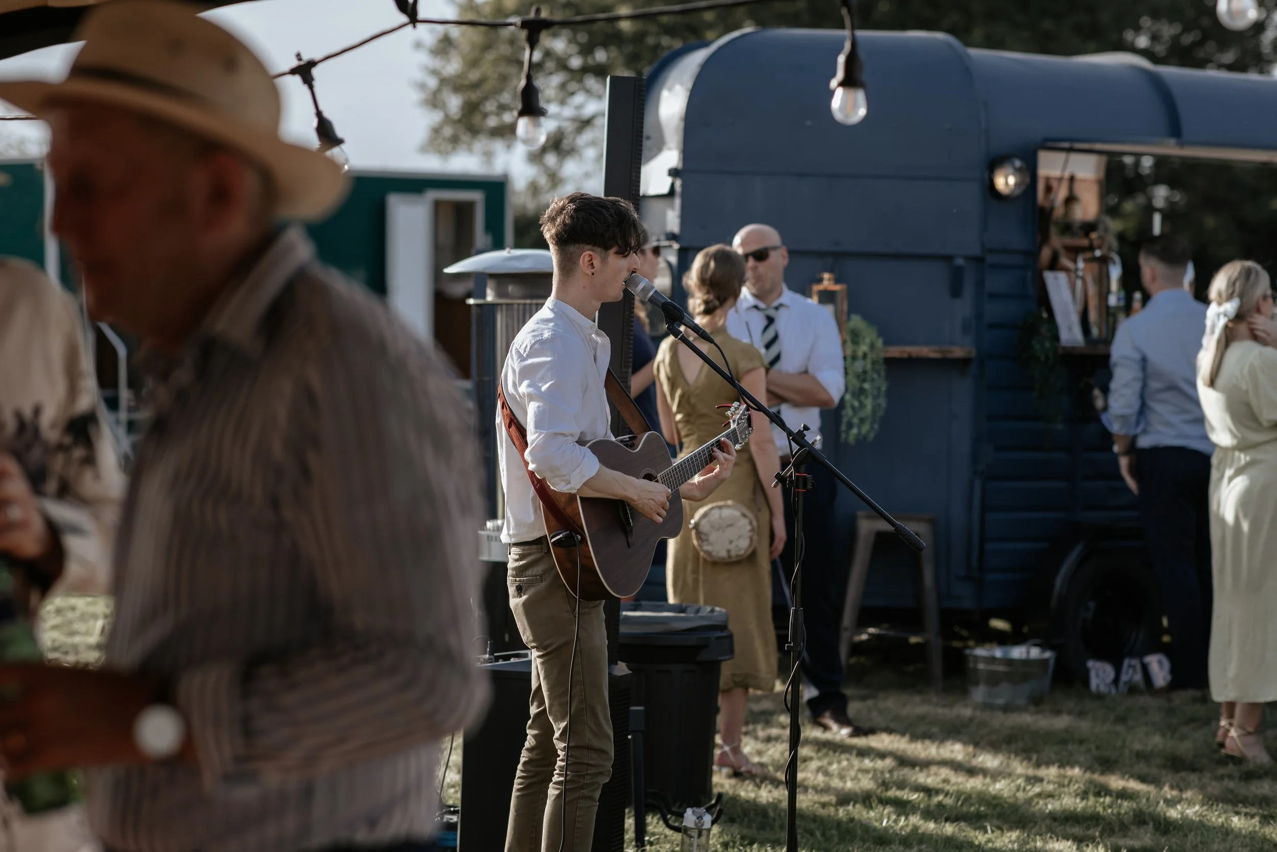 Young male  wedding musician playing an acoustic guitar and singing into a microphone at an outddoor wedding, with guests mingling and a food truck in the background.