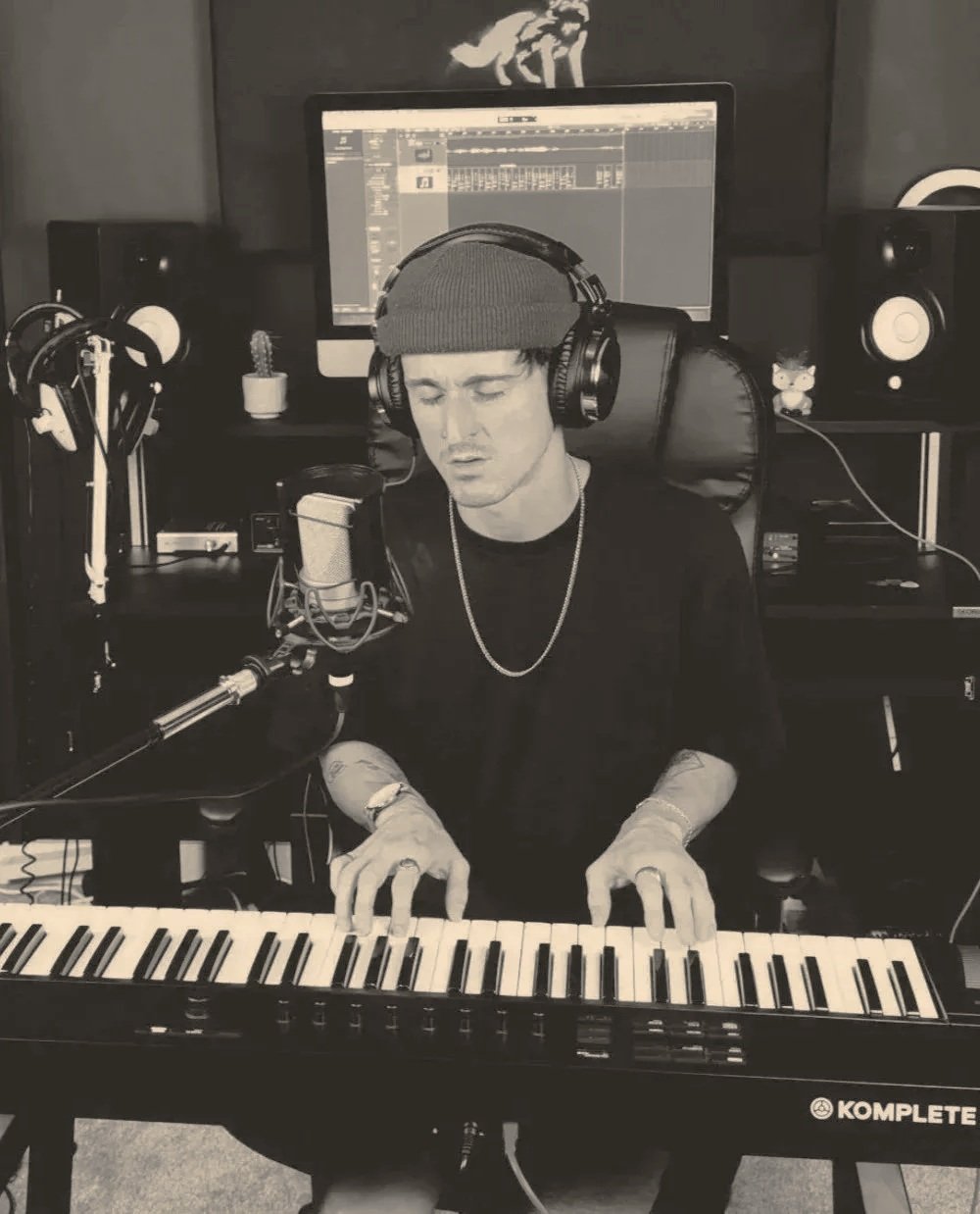A male musician wearing a beanie and headphones, singing into a microphone while playing a keyboard in a recording studio.