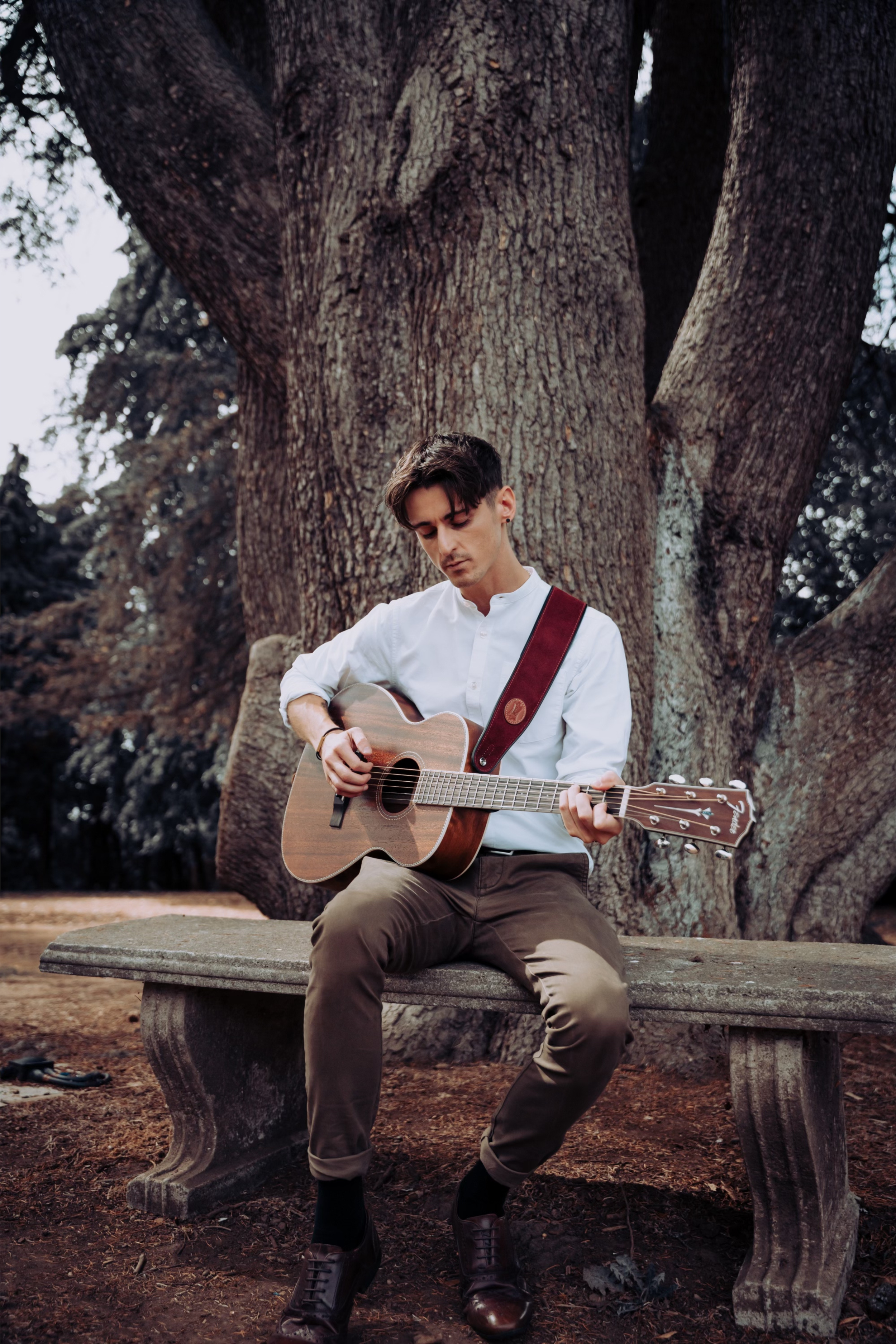 A young man with dark hair, dressed in a white shirt and brown pants, playing an acoustic guitar on a stone bench in a park with a large tree in the background.