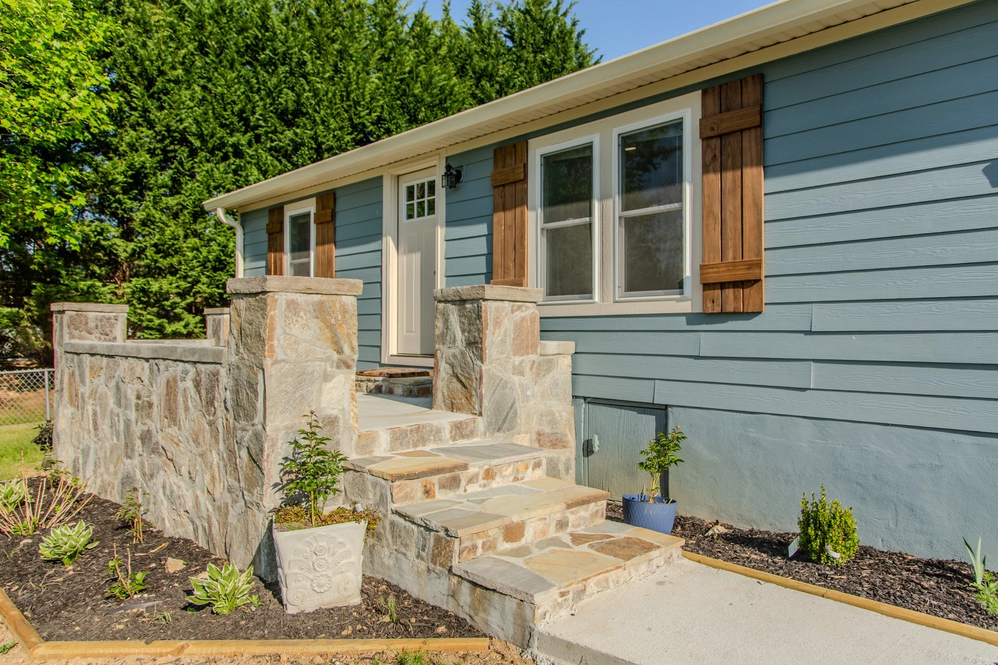 Front view of a blue house with wooden shutters, a small porch with stone steps, potted plants, and a garden bed with plants.