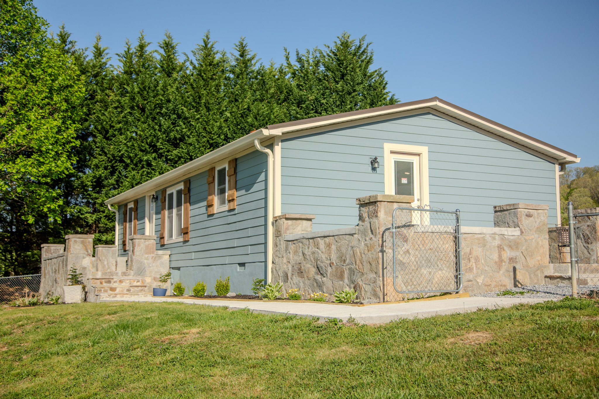 A blue house with stone accents, small front steps, and a chain-link gate, set in a green yard with trees in the background and a clear blue sky.