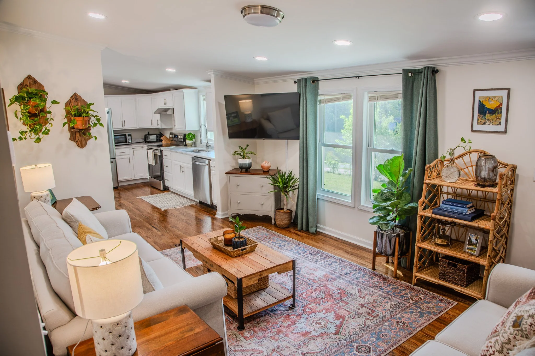 Living room with white couch, wooden coffee table, and green plants; open kitchen in background; large windows with green curtains.