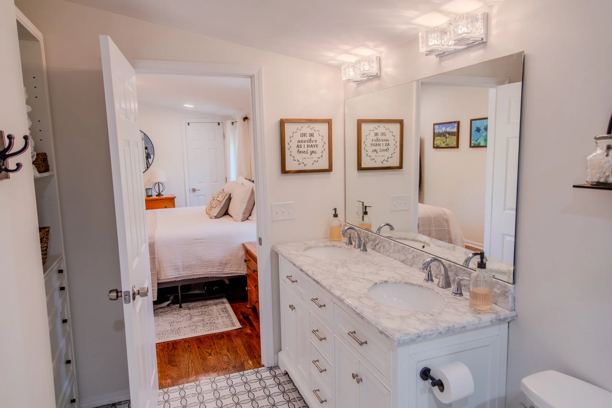 Bathroom with white vanity, marble countertop, double sink, large mirror, two wall-mounted light fixtures, framed wall art, soap dispenser, and view into bedroom with bed, pillows, and wooden nightstand.