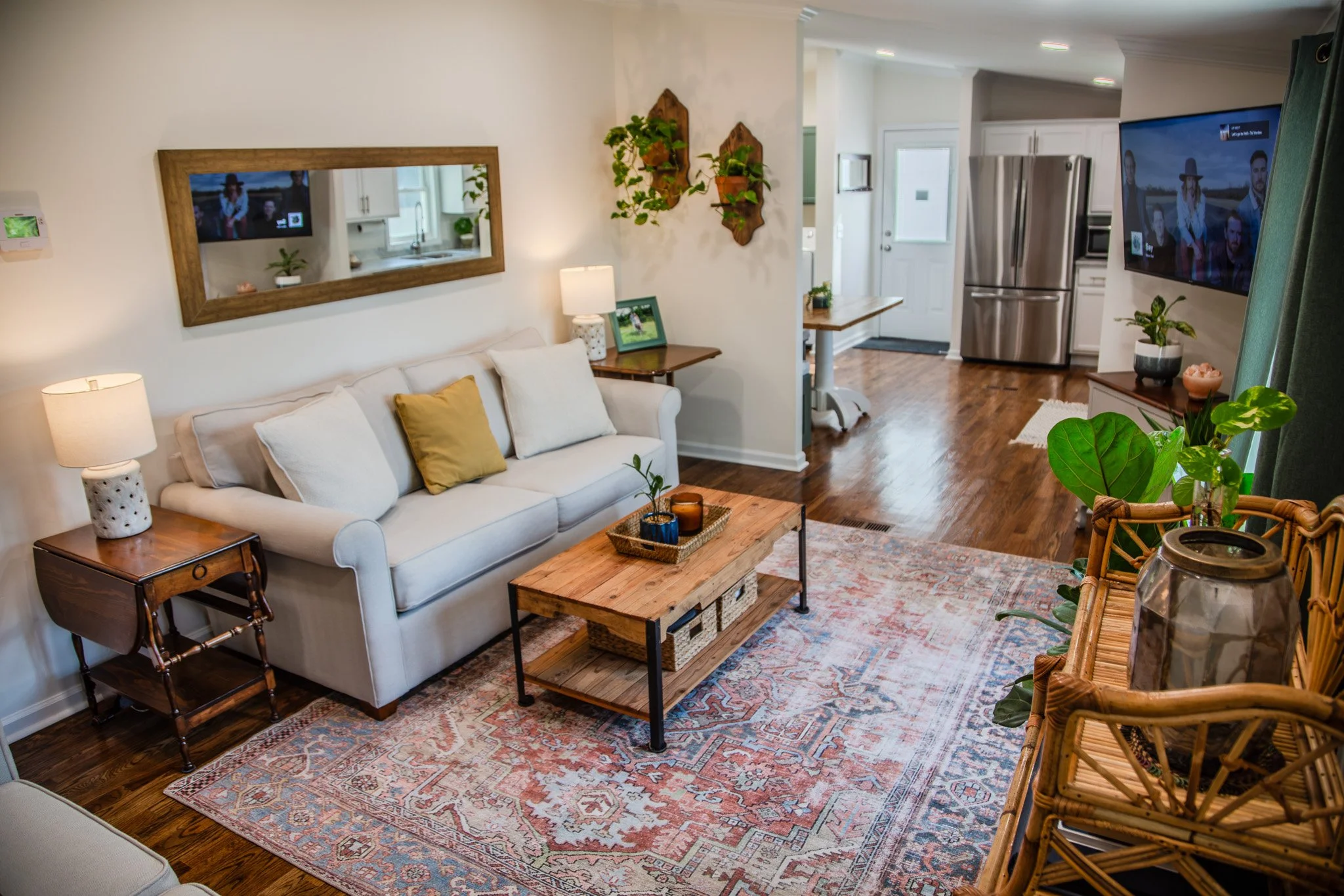 Cozy living room with white sofa, wooden coffee table, patterned area rug, and plant decor, connected to kitchen with stainless steel refrigerator.