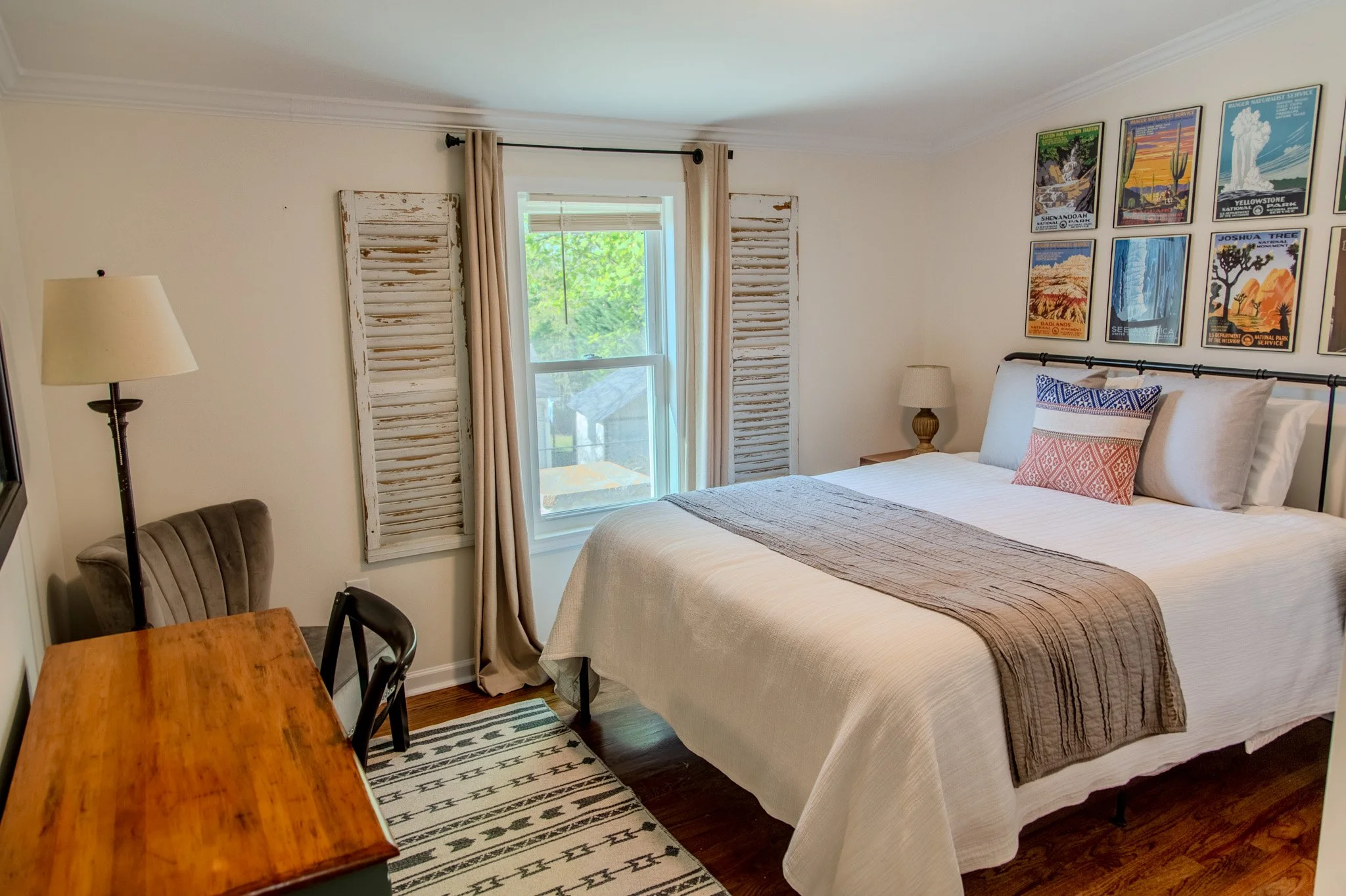 Cozy bedroom with a large bed, decorated with pillows and a brown bed runner, a wooden desk with a gray chair, a standing lamp, artwork on the wall, and a window with beige curtains and distressed shutters.
