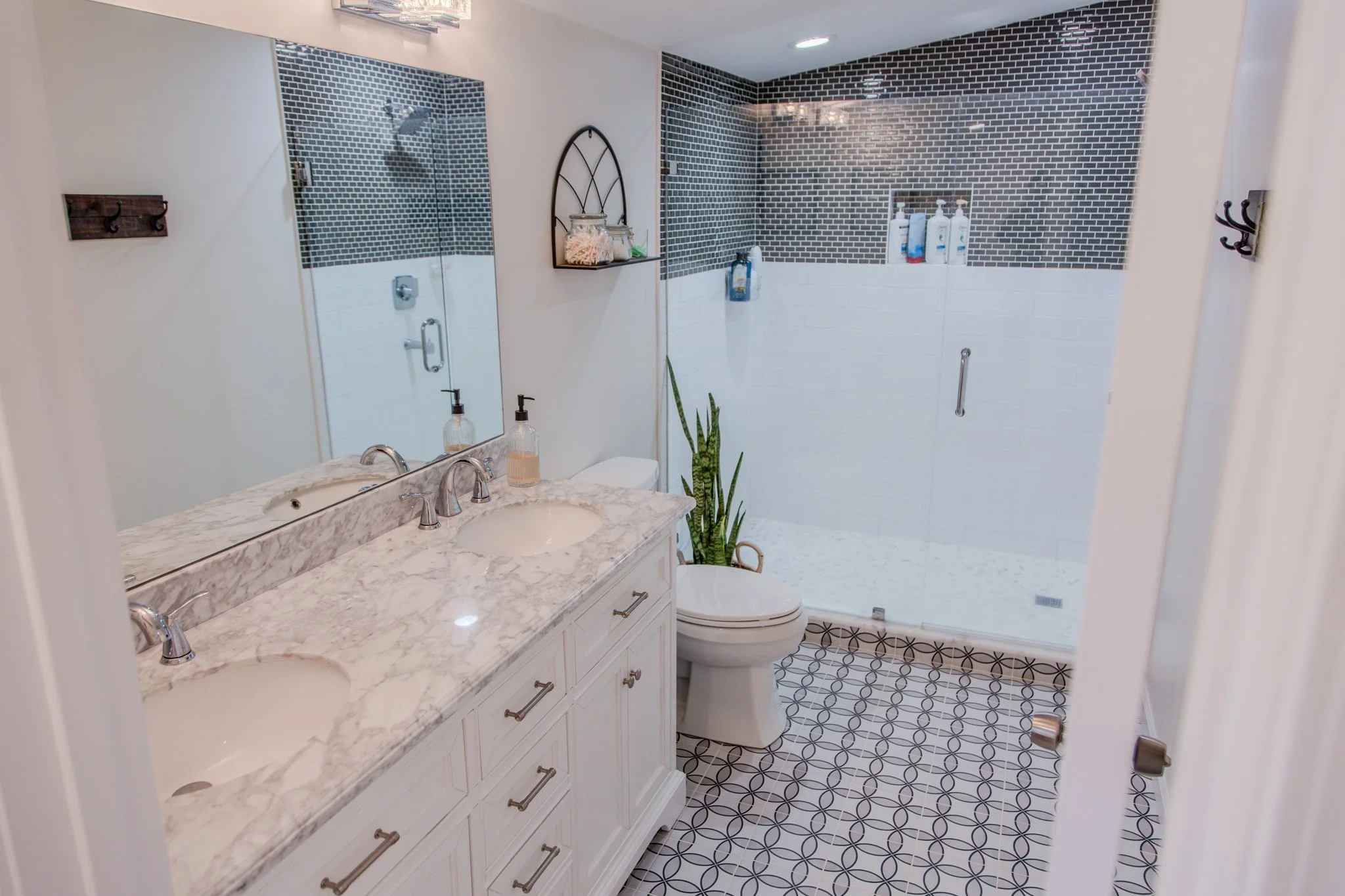 A bathroom with a double sink vanity with a marble countertop, a mirror, a toilet, and a glass-enclosed shower with dark mosaic tiles on the upper half and white tiles on the lower half. There is a plant next to the toilet and wall hooks on the side walls.