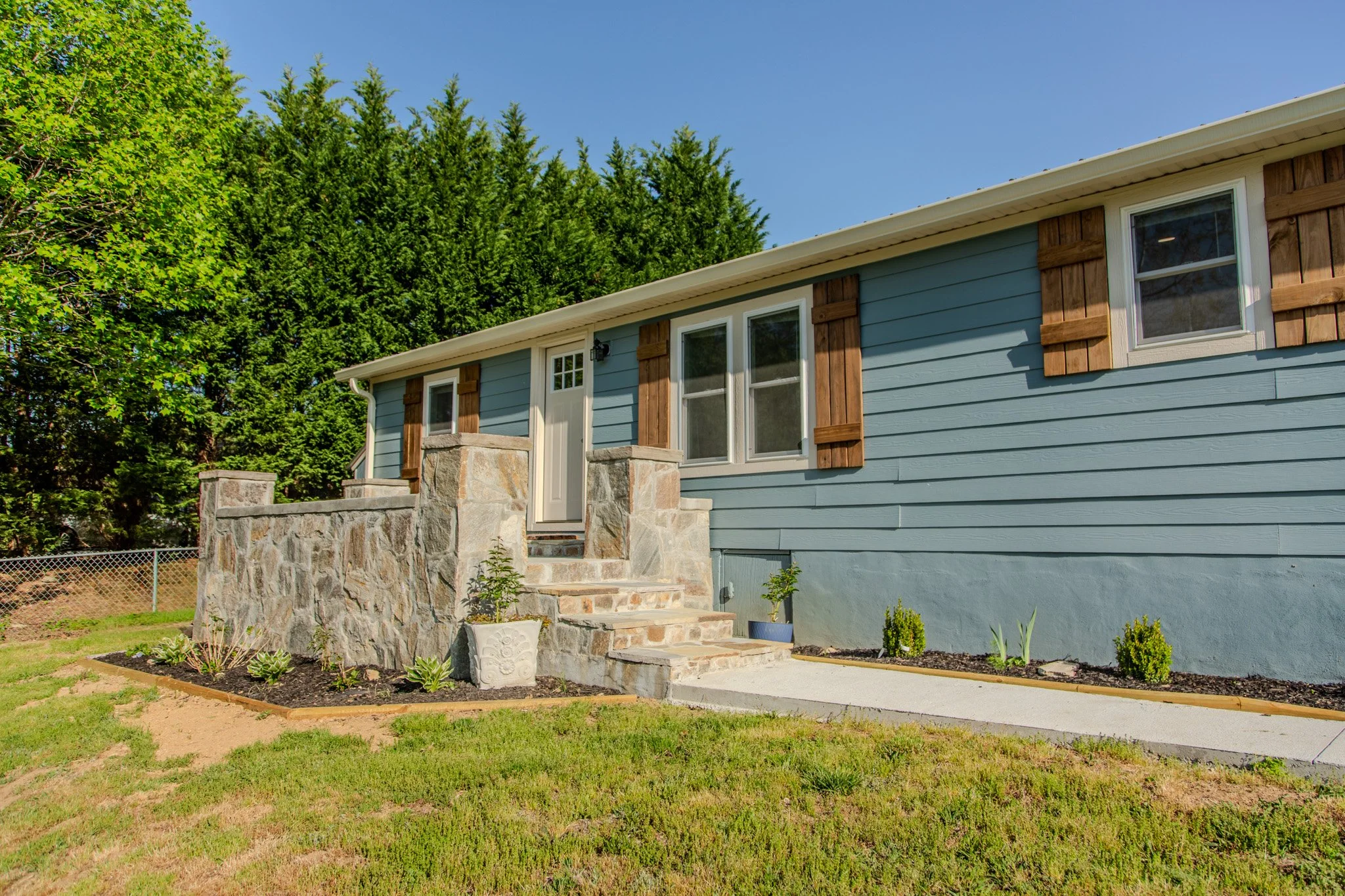 Side view of a blue house with stone stairs and a white door, surrounded by green trees and a lawn with small plants.