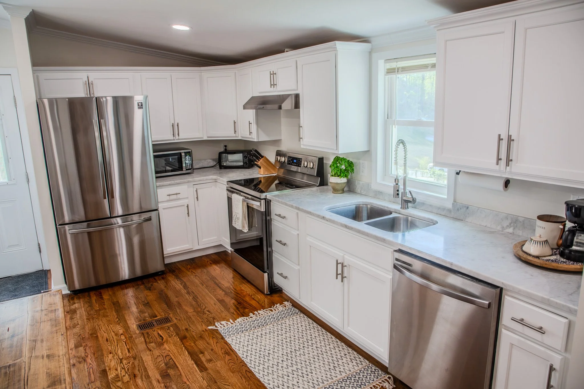 Modern kitchen with white cabinets, stainless steel appliances, marble countertops, wooden flooring, and a window above the sink.