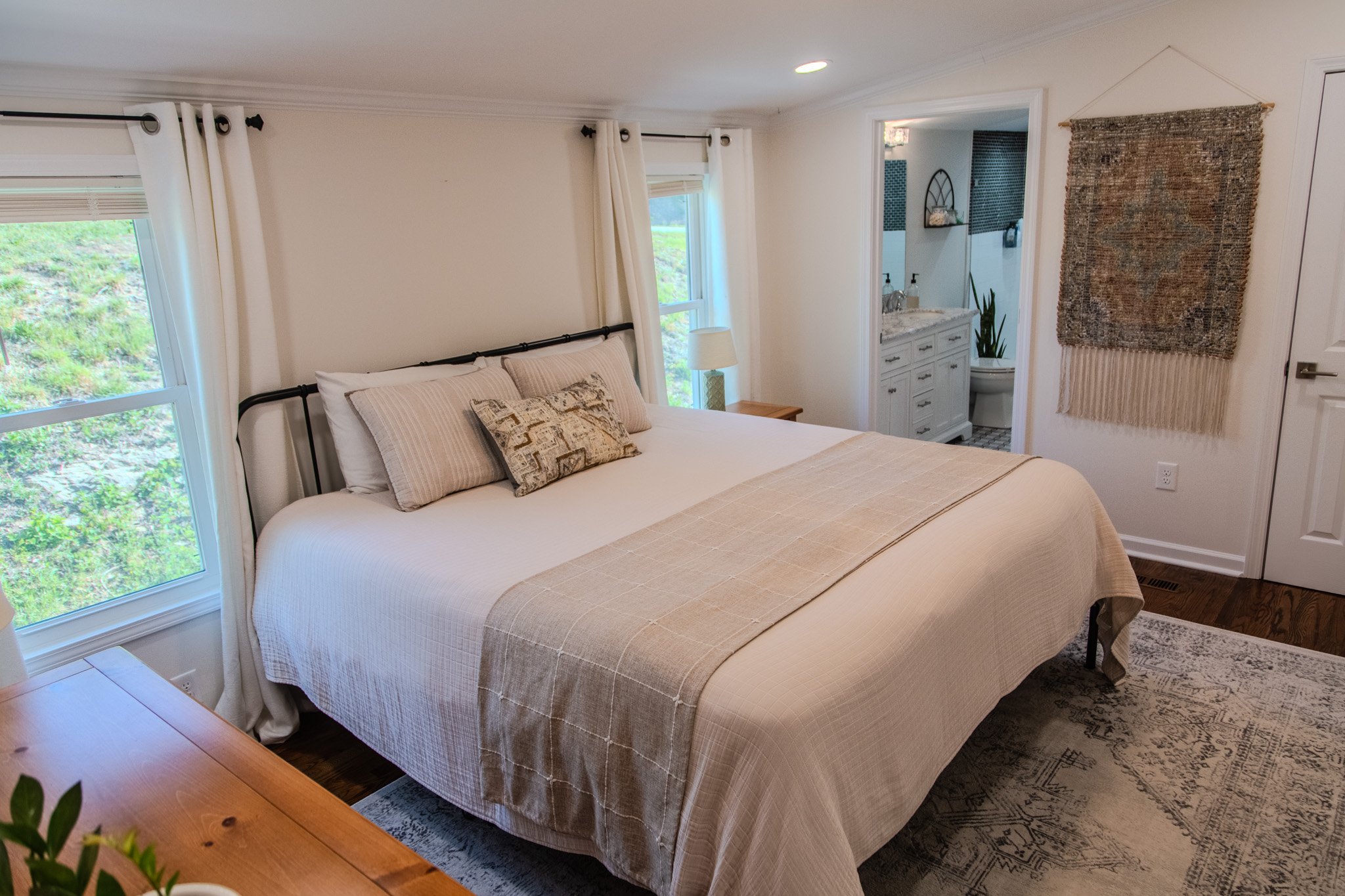 Modern bedroom with a king-sized bed, cream bedding, and decorative pillows, next to windows with white curtains, adjacent to an en-suite bathroom with a vanity, mirror, and toilet, and a wall hanging on the right side of the wall.