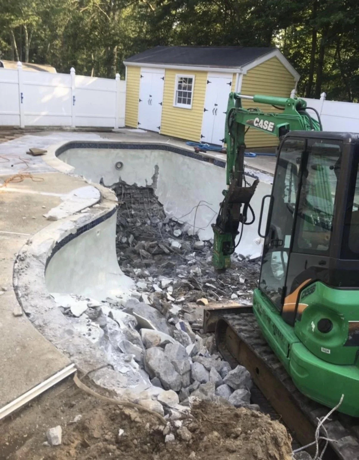 A backyard pool undergoing renovation or demolition, with a small yellow shed in the background and construction equipment, including a green mini excavator, inside the pool area. The pool is partially torn down, showing debris and construction material.