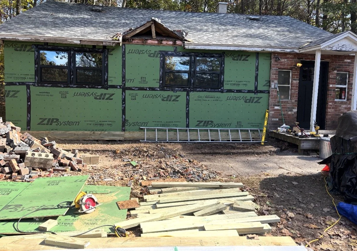 Front of a house under renovation, with siding partially removed, construction tools and materials on the ground, and windows installed in the front wall.