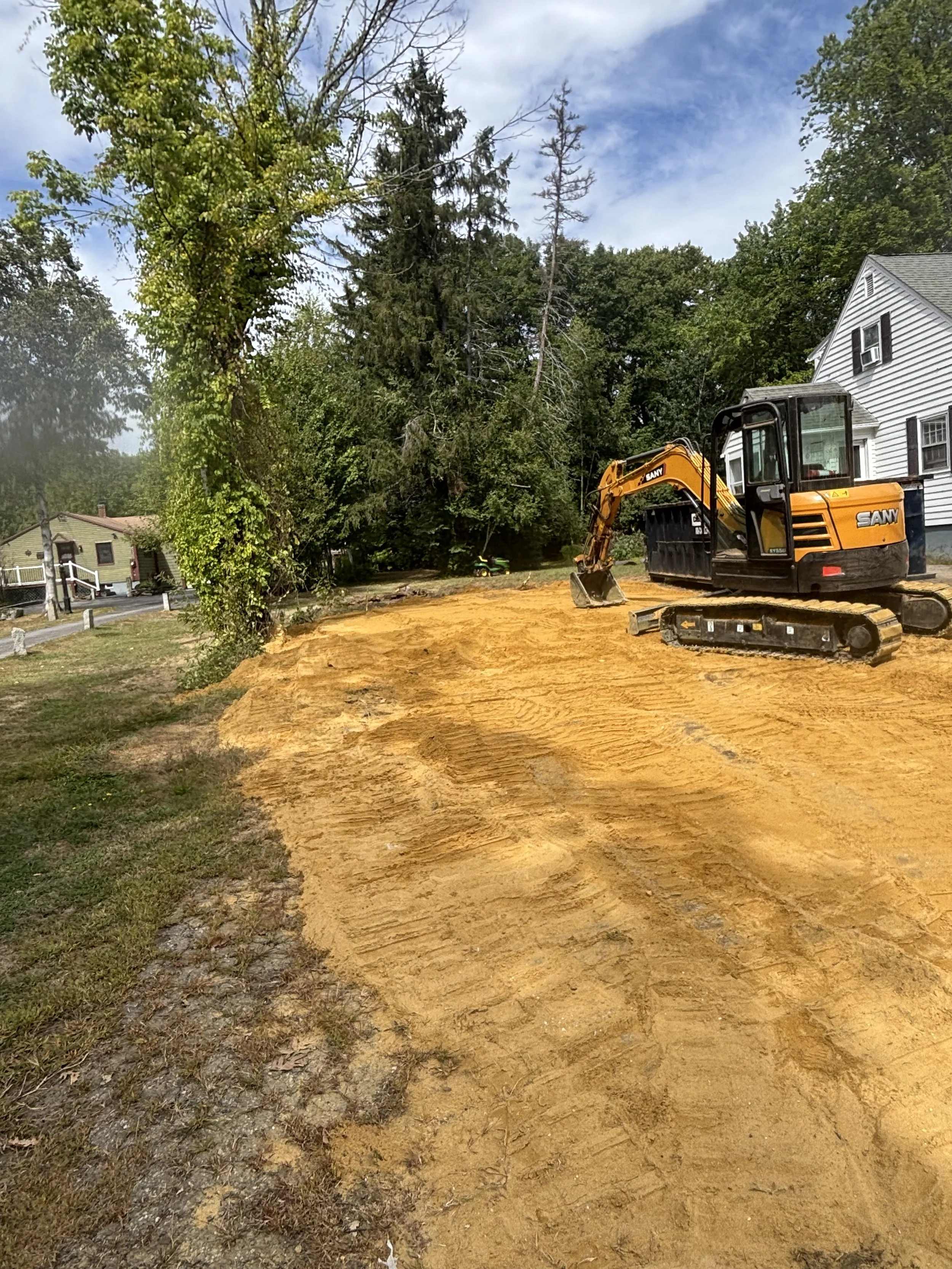 A small excavator parked on a sandy patch of land next to a white house, with trees and a partly cloudy sky in the background.