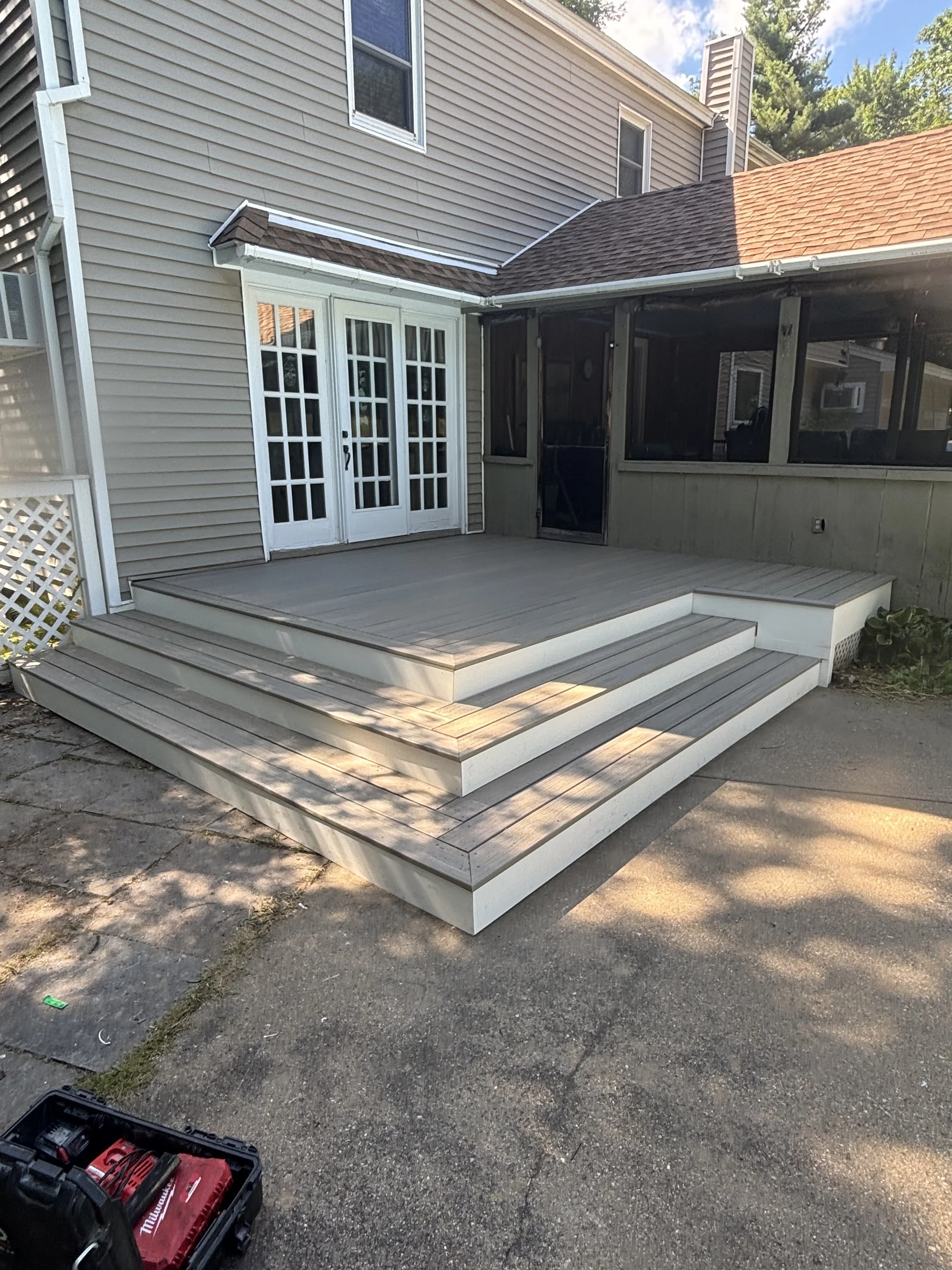 A backyard wooden deck with four steps leading up to the house, attached to a beige house with a sliding glass door and screened porch, under a partly cloudy sky.