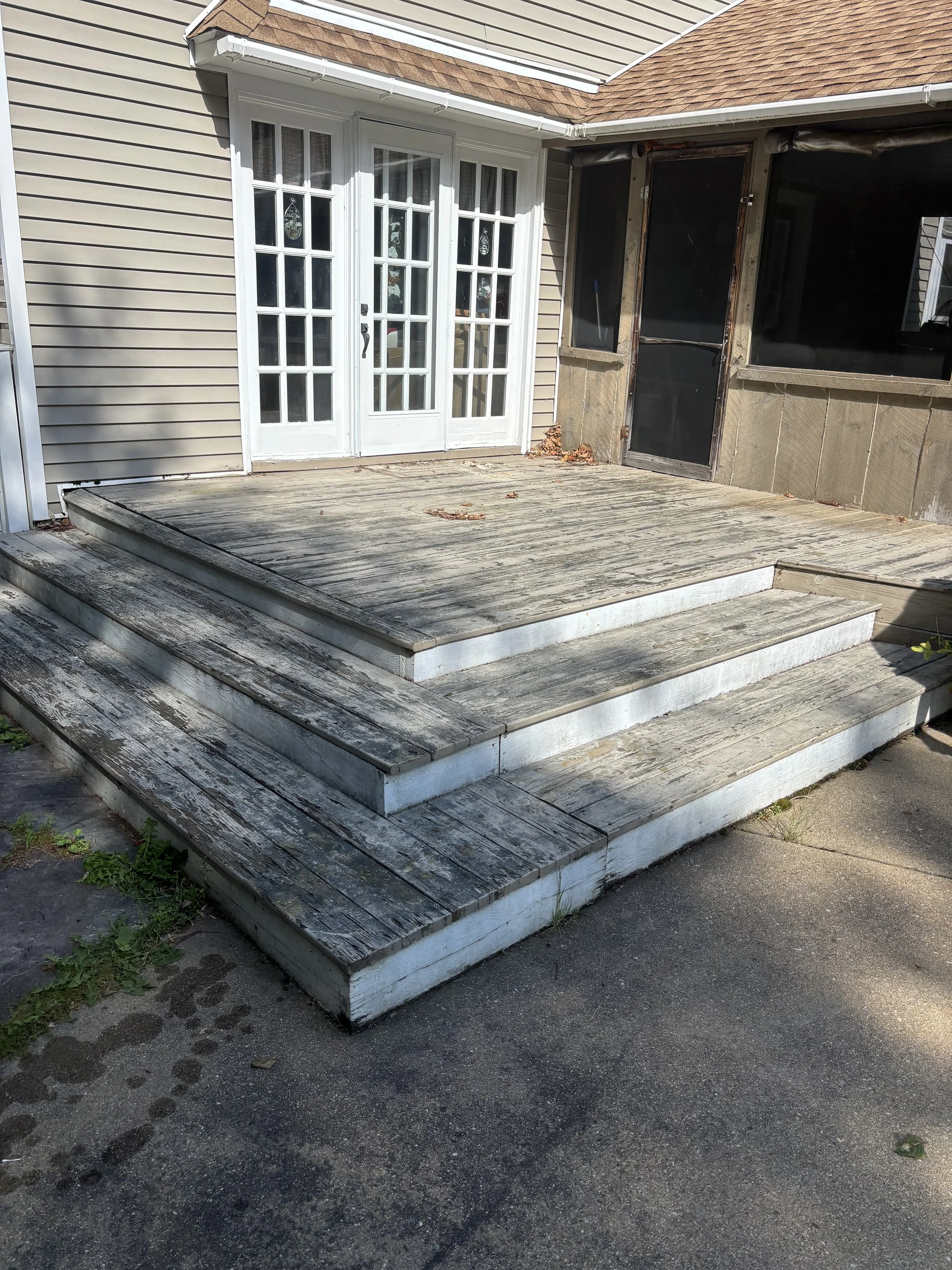 A weathered wooden deck with three steps leading up to double glass doors on a house. The house has beige siding and a screened porch on the right side. Some leaves are scattered on the deck.