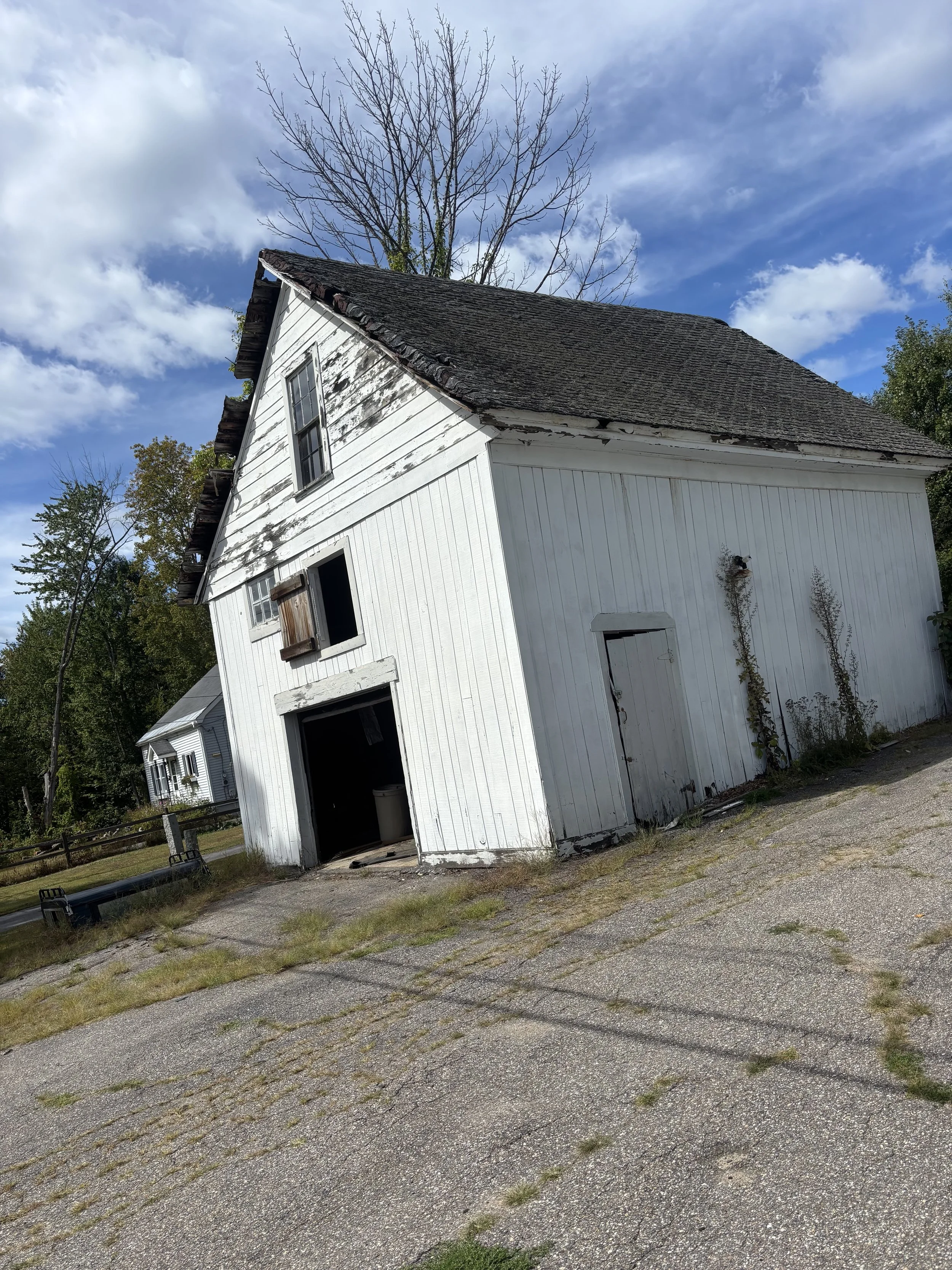 An old white barn with peeling paint, a small window with shutters, and a large open door showing the inside. The barn has a steeply pitched roof covered in dark shingles. There are dry weeds growing at the base and a weathered smaller door on the side. The sky is partly cloudy with patches of blue, and a few trees are visible in the background.