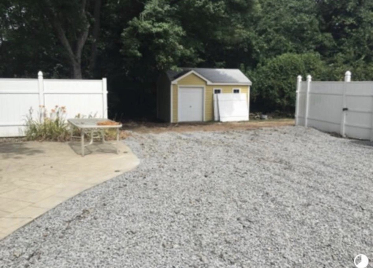 Backyard with gravel ground, white fence, a small yellow shed, a bench, and plants.