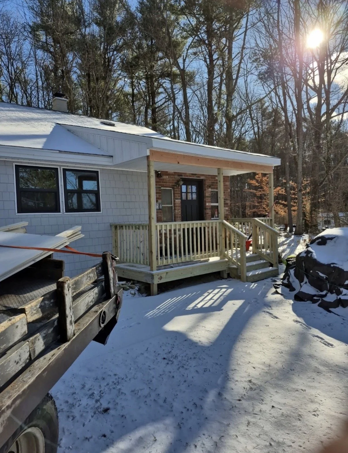A house with a small wooden porch under construction, surrounded by snow on the ground and trees in the background, with sunlight shining through.