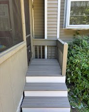 Small gray staircase with three steps leading up to a porch or doorway, next to a window and a garden with green plants.