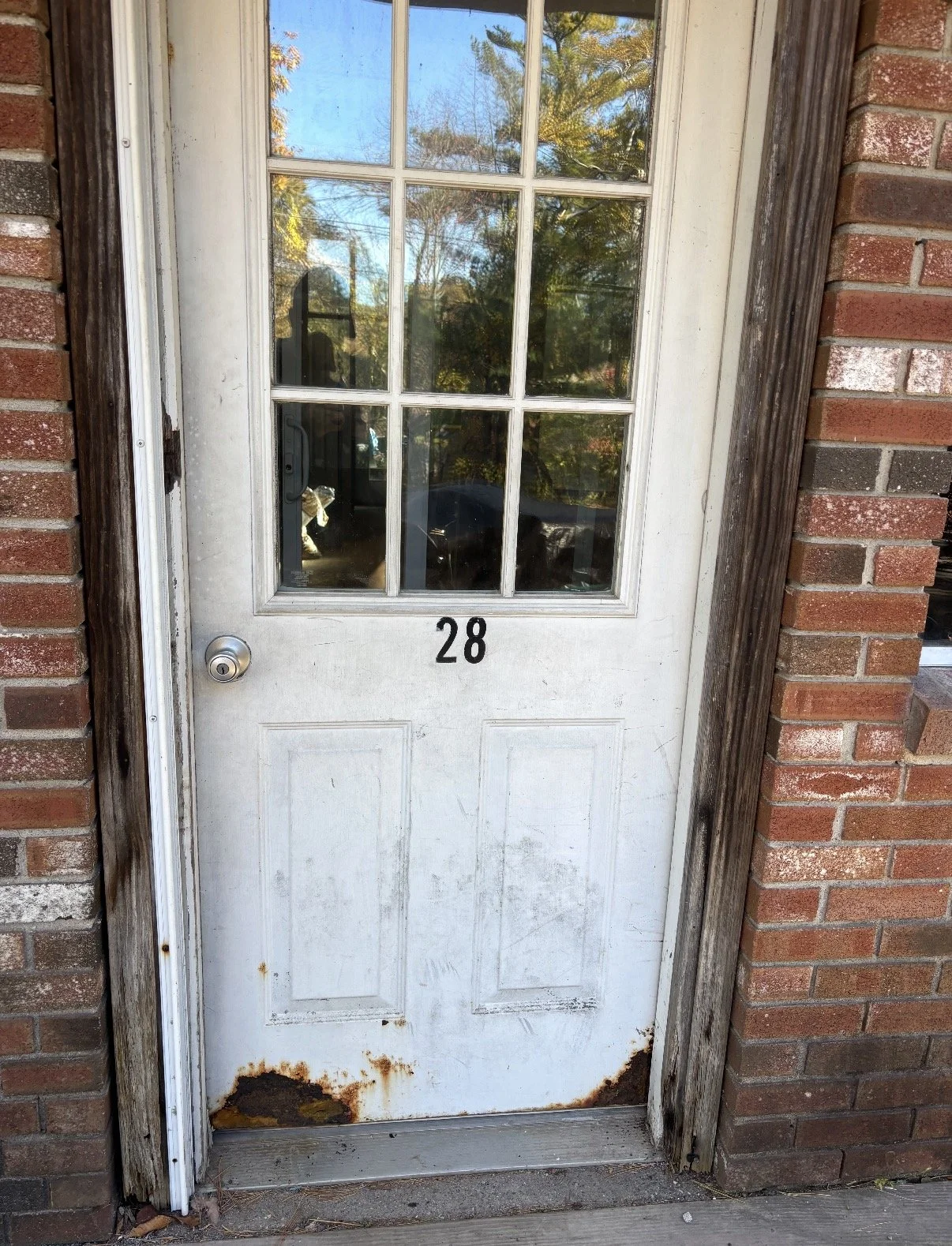 A white front door with a window and the number 28 below it, surrounded by weathered brick and wood framing, with rust and dirt at the bottom of the door.