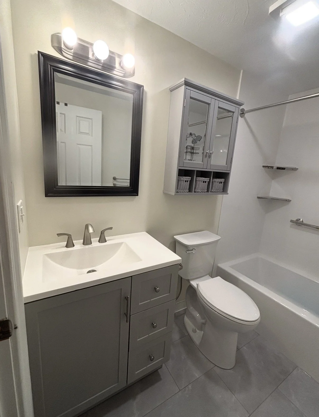 A small modern bathroom with a white sink vanity, a framed mirror, a gray wall-mounted cabinet with glass doors, a white toilet, a bathtub with a shower curtain rod, and beige tiled flooring.