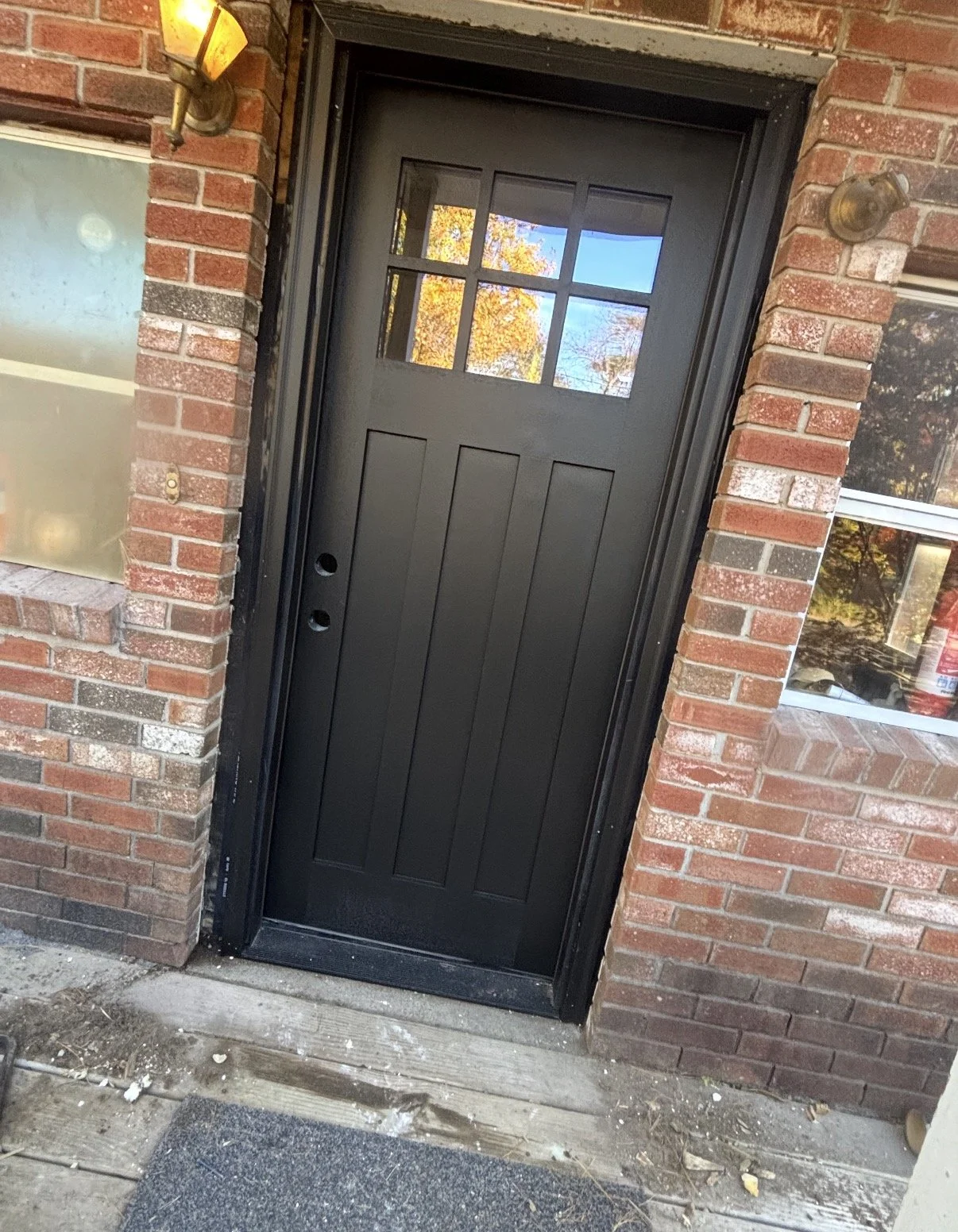 Black front door with a window at the top, surrounded by brick walls, with outdoor wall lights on either side.
