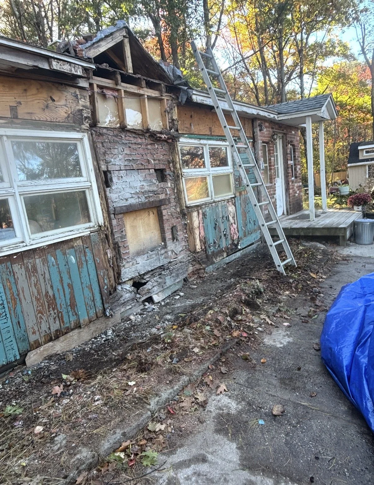 The image shows a house undergoing repair or demolition, with the exterior wall partially torn down, exposing the structure inside. A ladder leans against the house, and the yard has some debris, leaves, and a large blue tarp.