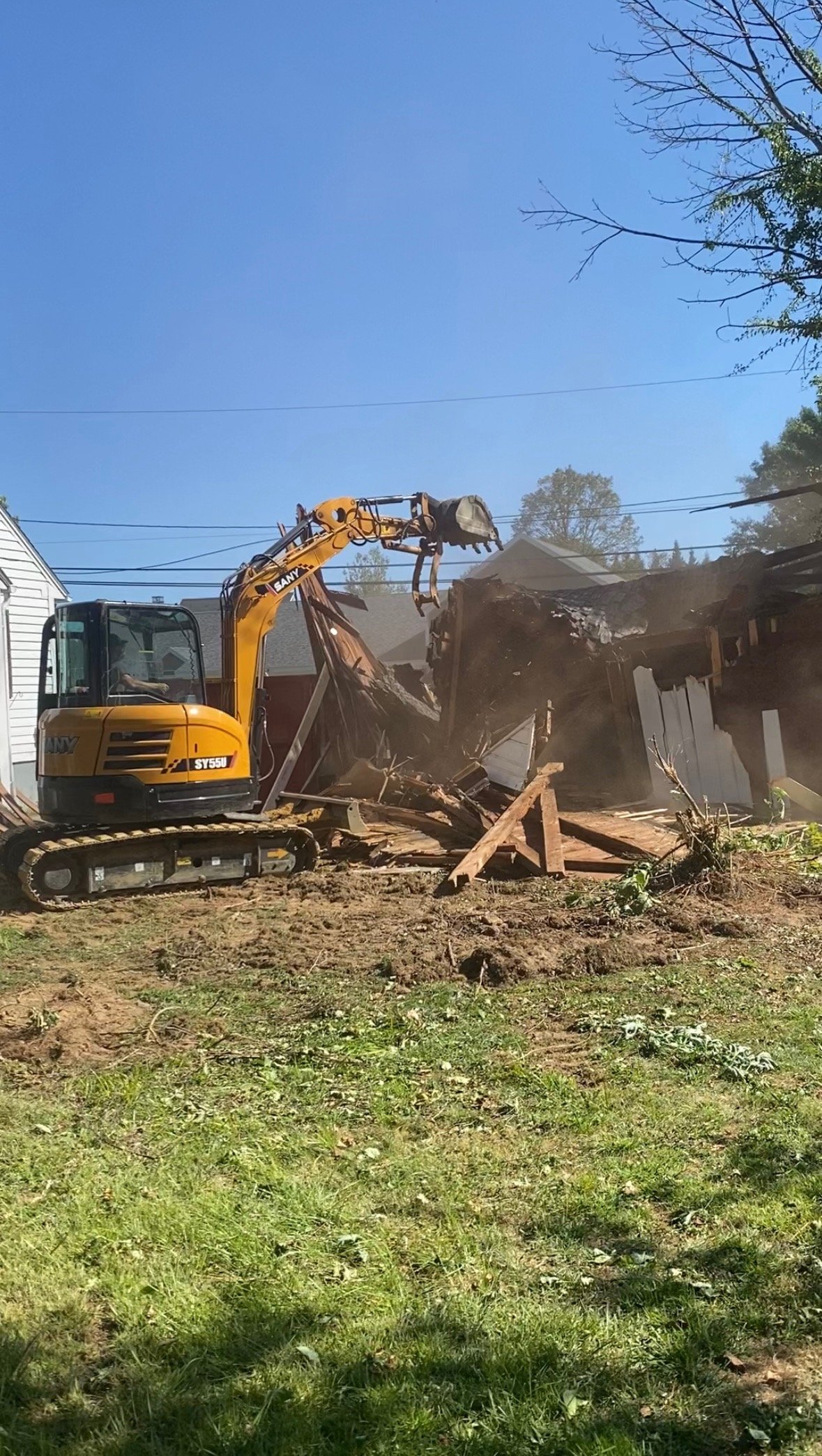 A yellow excavator demolishing a small wooden structure with a clear blue sky in the background.