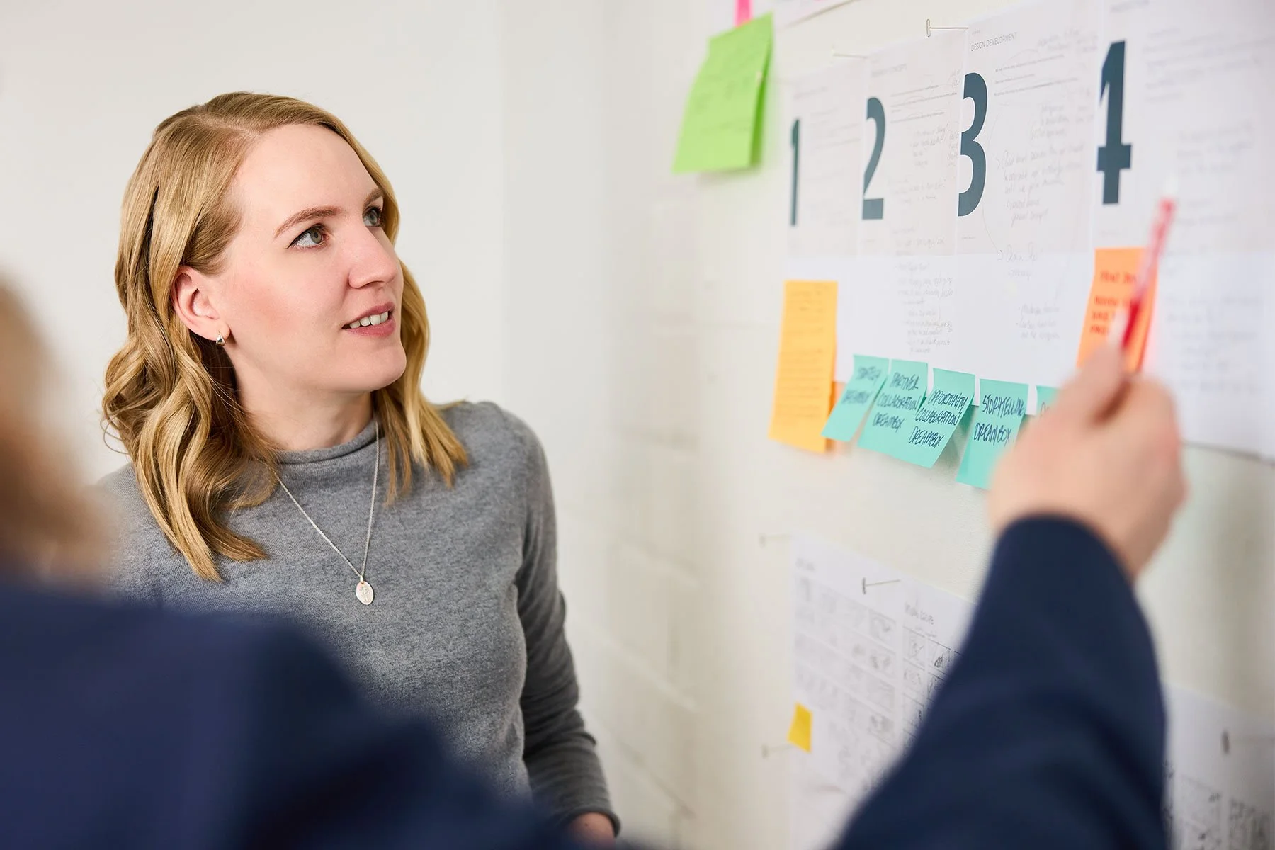 A blonde woman in a grey sweater looks thoughtfully at a wall of project phases.