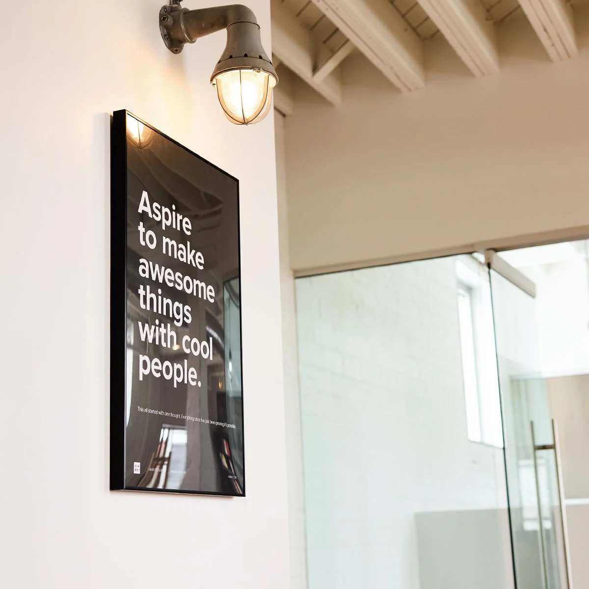 A framed black poster with white text that reads "Aspire to make awesome things with cool people" hangs on a white wall beneath an industrial-style light fixture.