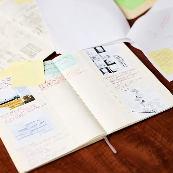 A top-down shot of two open notebooks on a wooden table, filled with architectural sketches, project notes, and pasted-in diagrams.