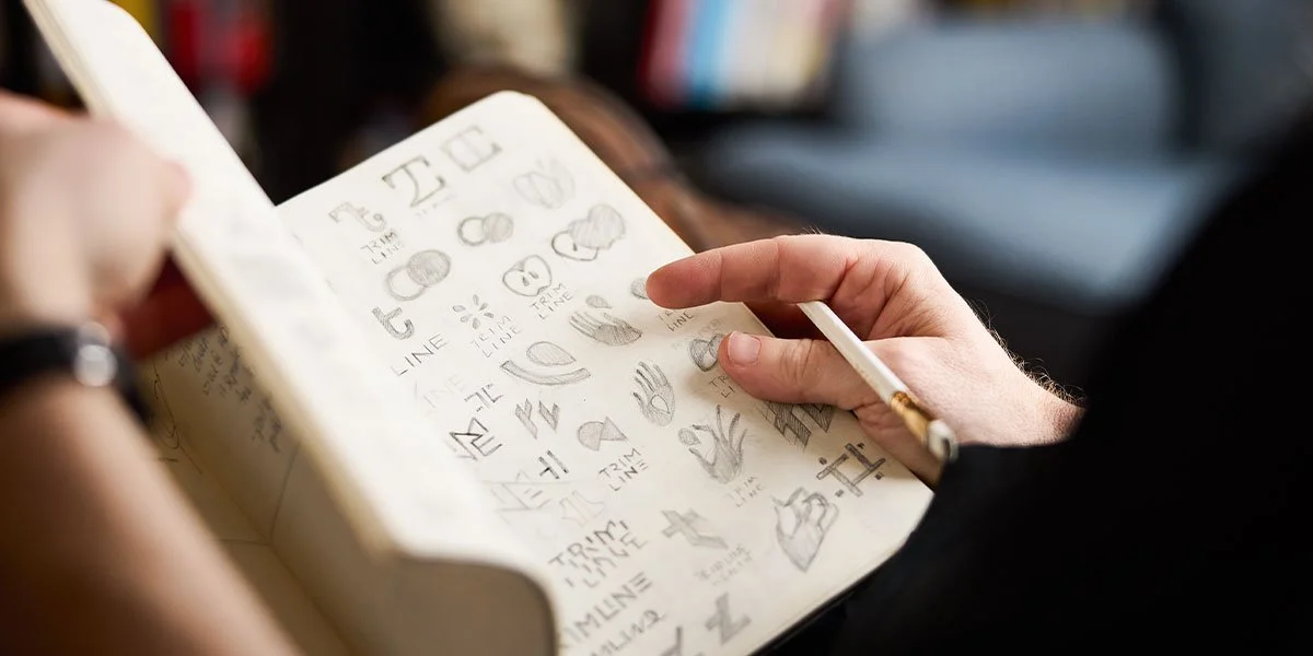 A close-up, shallow-focus shot of a person’s hands holding an open sketchbook filled with various pencil-drawn logo concepts and icons, including hand shapes and typography.